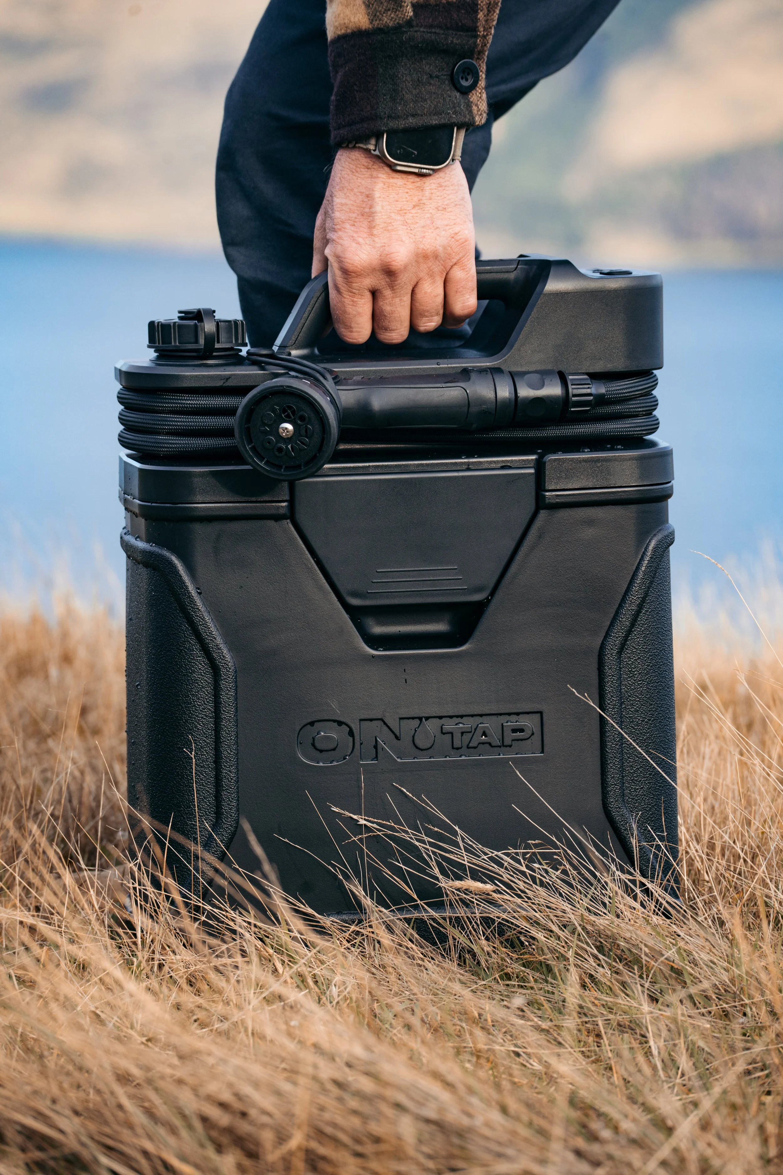 Close-up of a person holding a portable black device with a handle and a coiled cable, standing outdoors in tall grassy field.