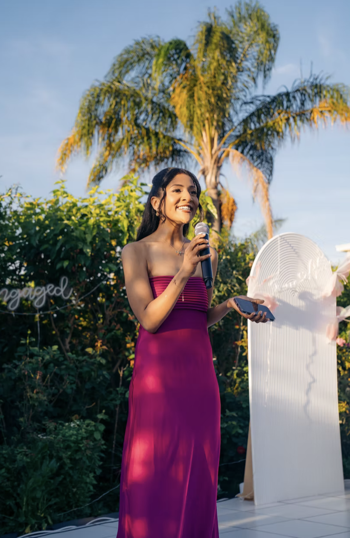 A woman in a strapless magenta dress holding a microphone and a phone, standing outdoors near a palm tree with a clear blue sky in the background.