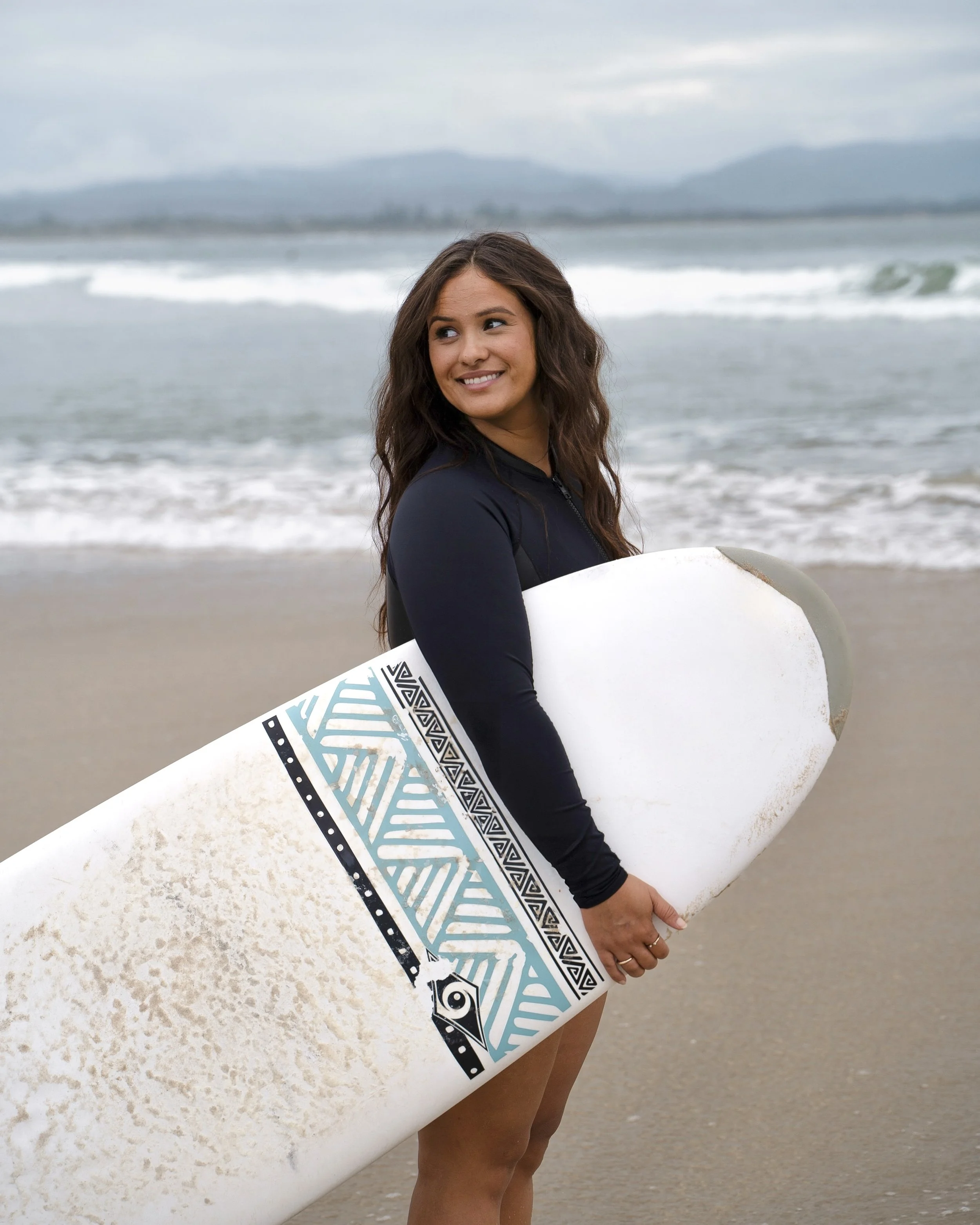Woman standing on the beach holding a surfboard, wearing a black wetsuit, with ocean waves and cloudy sky in the background.