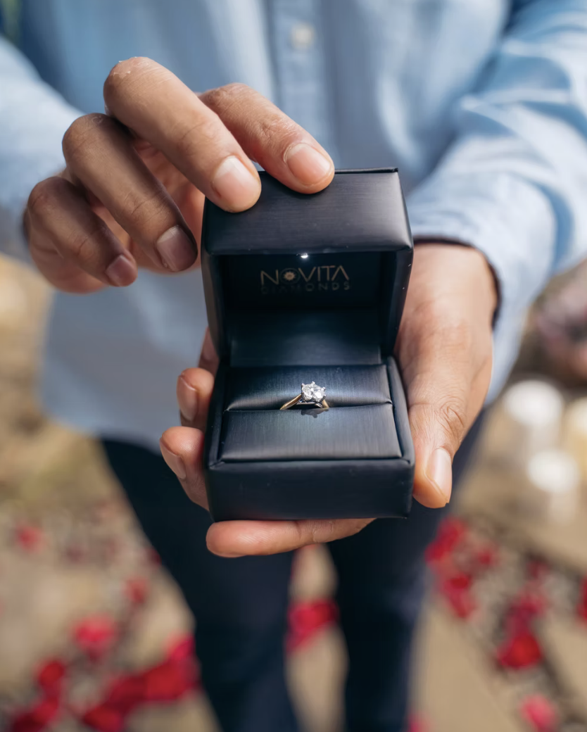 Person holding an open black jewelry box with a diamond engagement ring inside