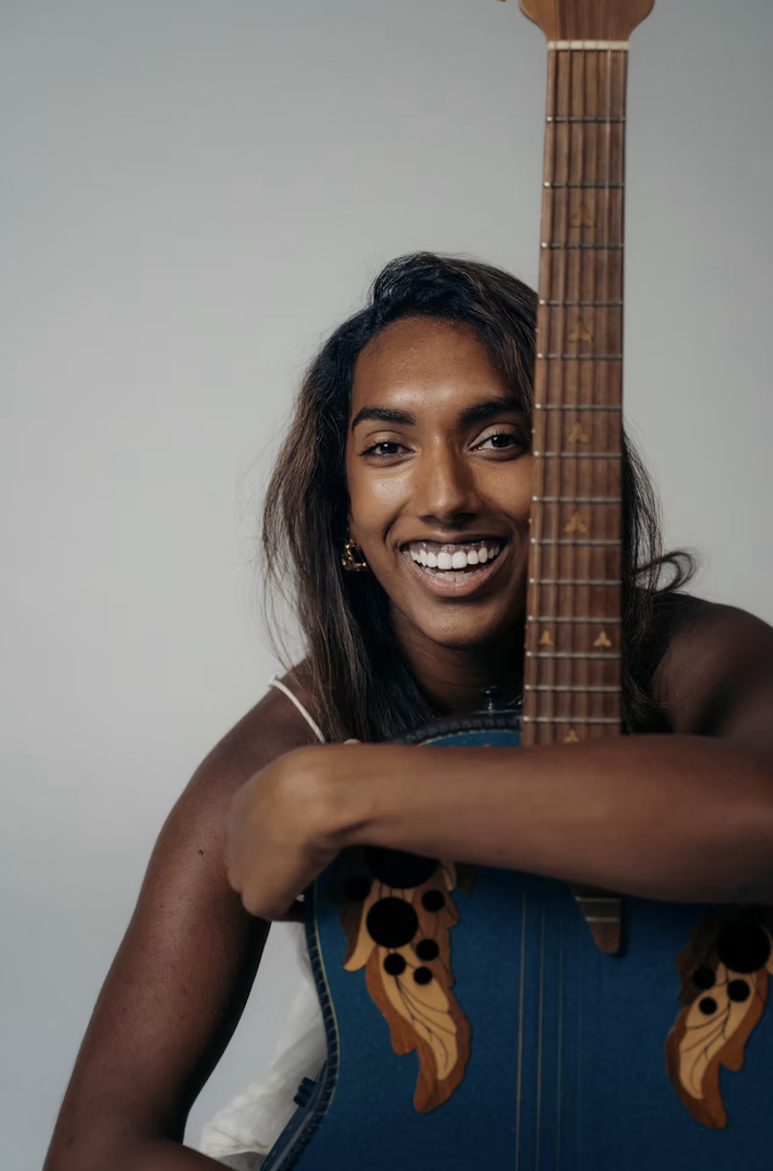 Smiling woman with dark hair holding an acoustic guitar in front of a plain light gray background.
