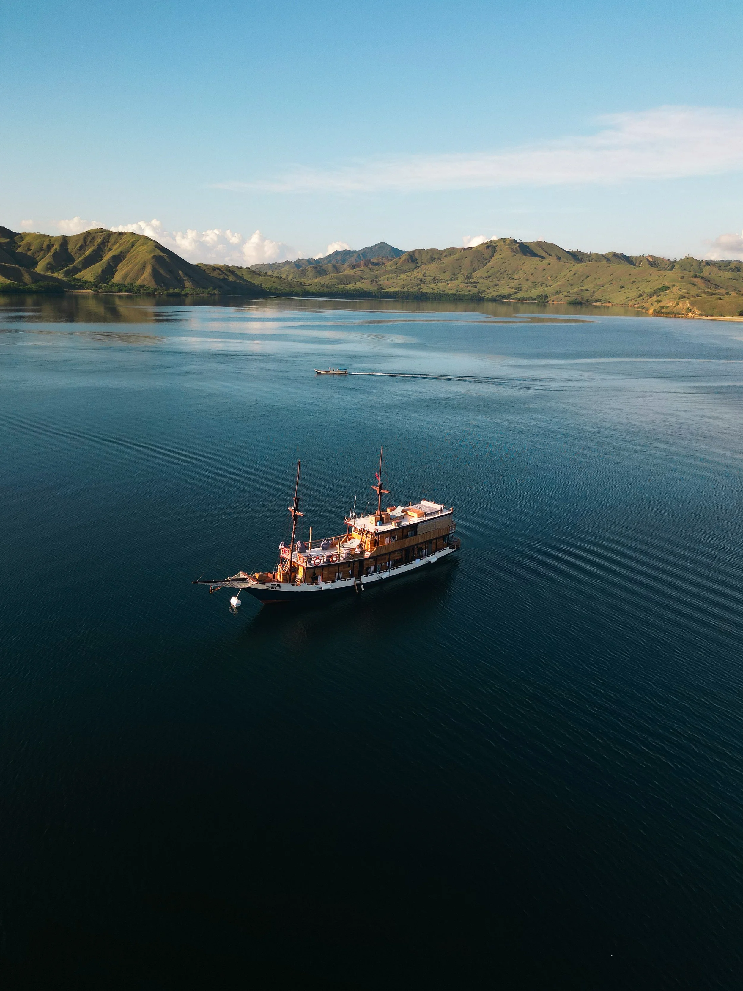 A sailboat on a calm lake with green hills and mountains in the background under a clear sky.