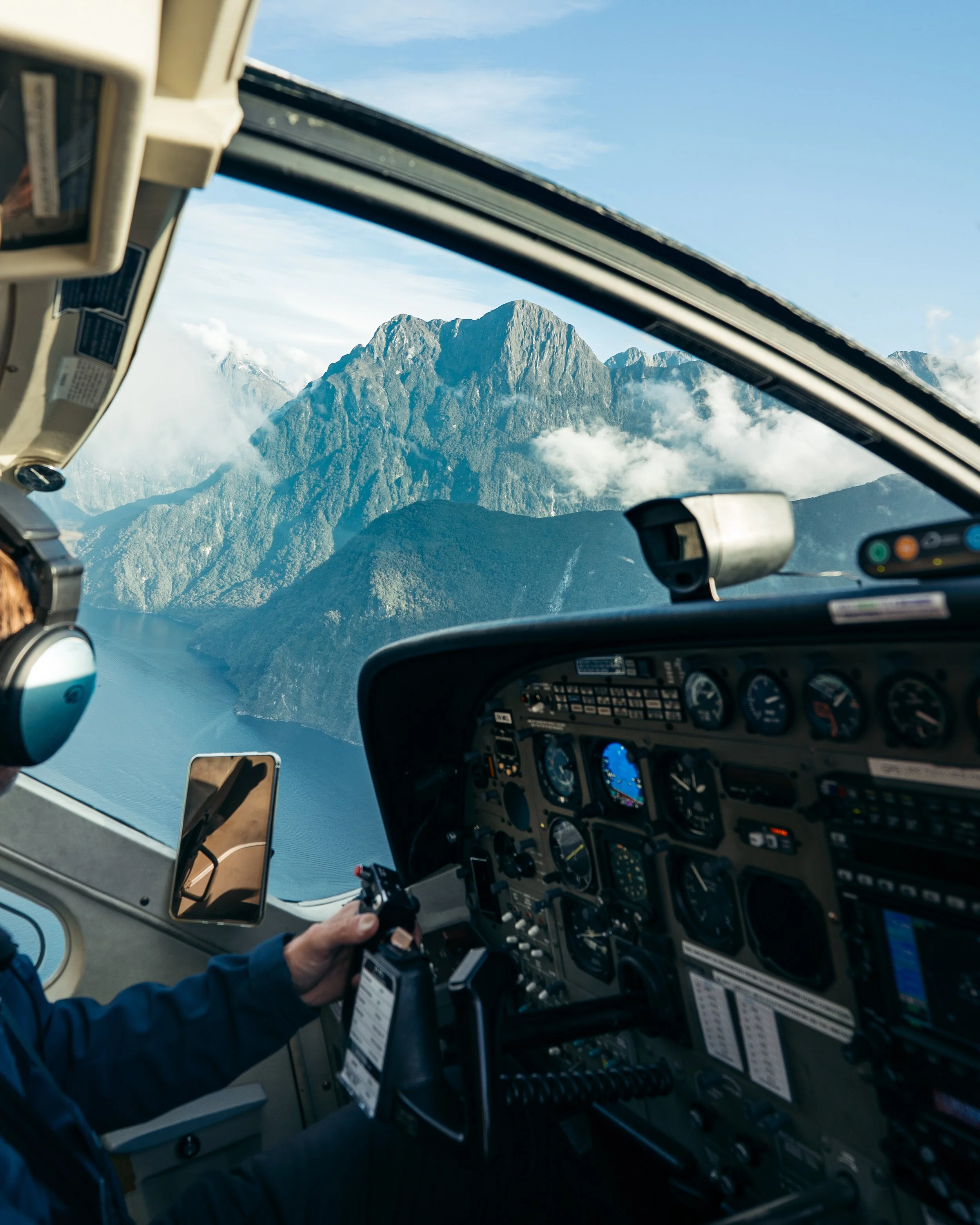 Inside the cockpit of an aircraft flying over mountains and clouds, with a view of the instrument panel and a pilot holding a device in their left hand.
