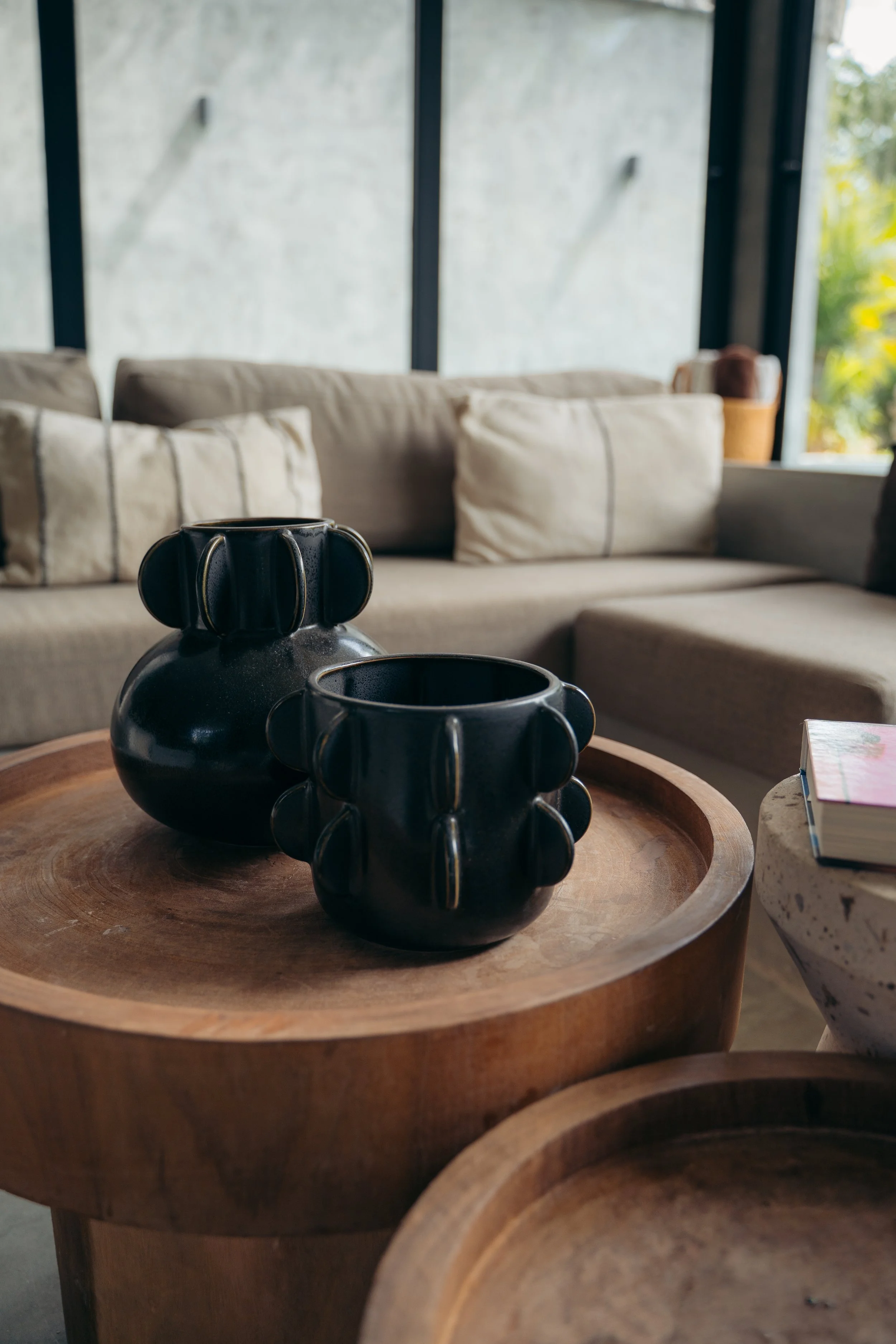 Close-up of two black ceramic vases on a round wooden table in a living room with beige couch and large windows.