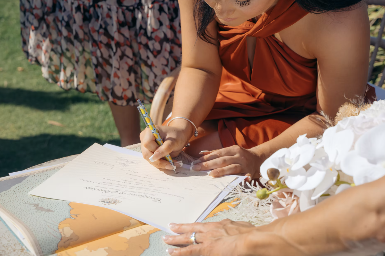 A woman in an orange dress signs a document outdoors on a table, with white flowers nearby.
