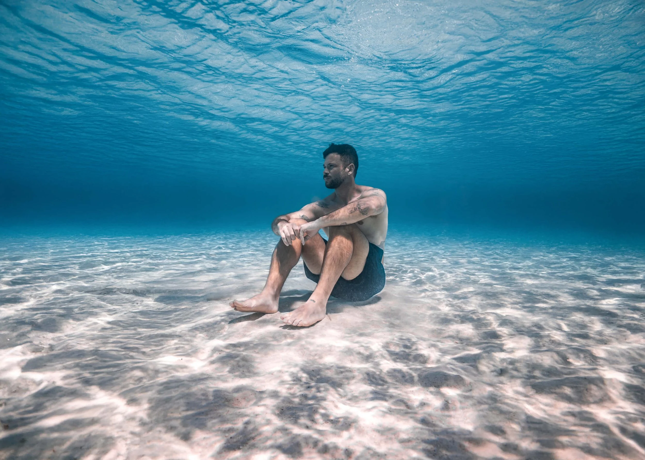 A man sitting on a beach underwater, with his arms wrapped around his knees, surrounded by clear blue water and ocean waves.