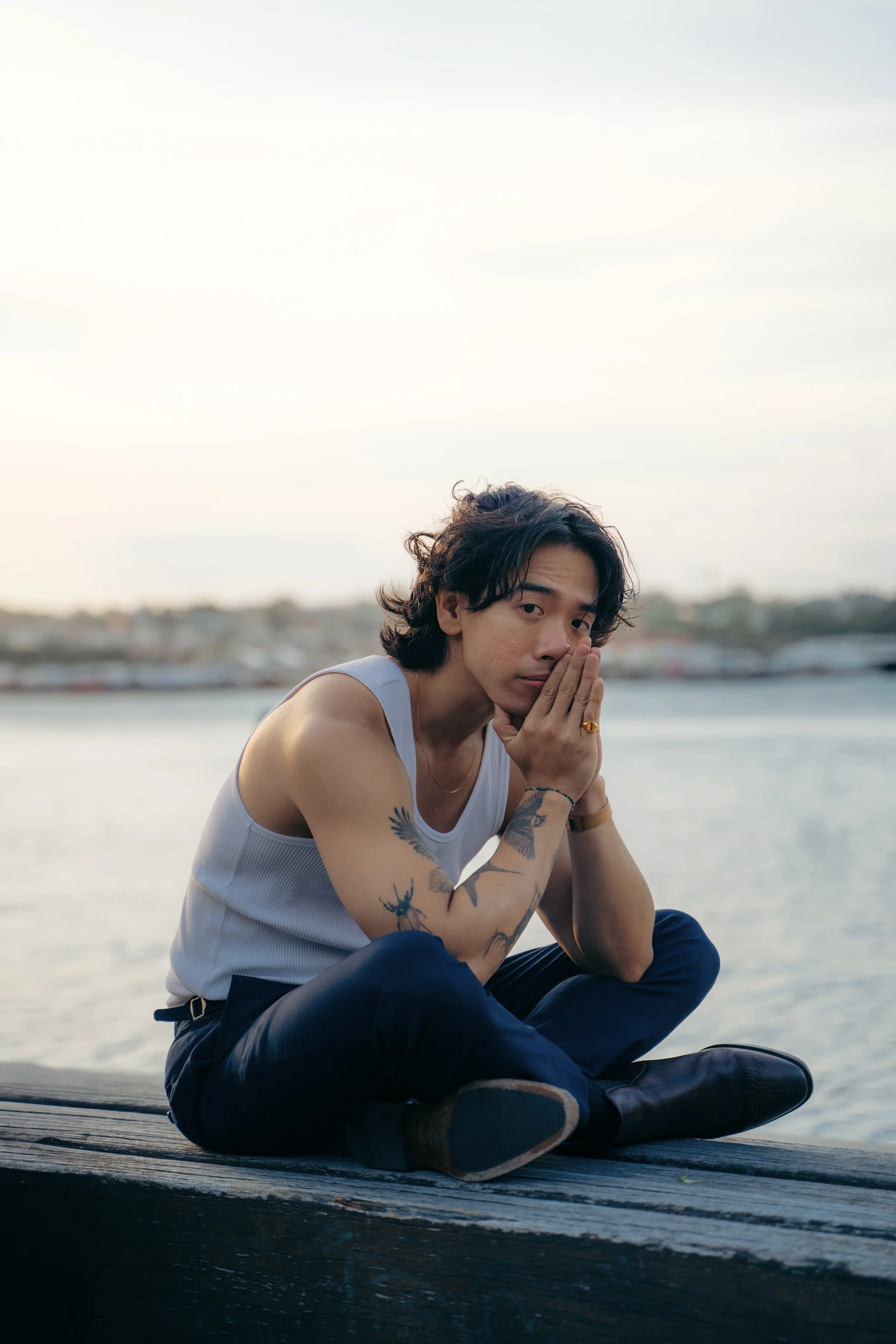 A young man with tattoos on his arms and chest, wearing a sleeveless shirt and dark pants, sitting cross-legged on a wooden dock near a body of water, with his hands pressed together in a prayer-like gesture, during evening or sunset.