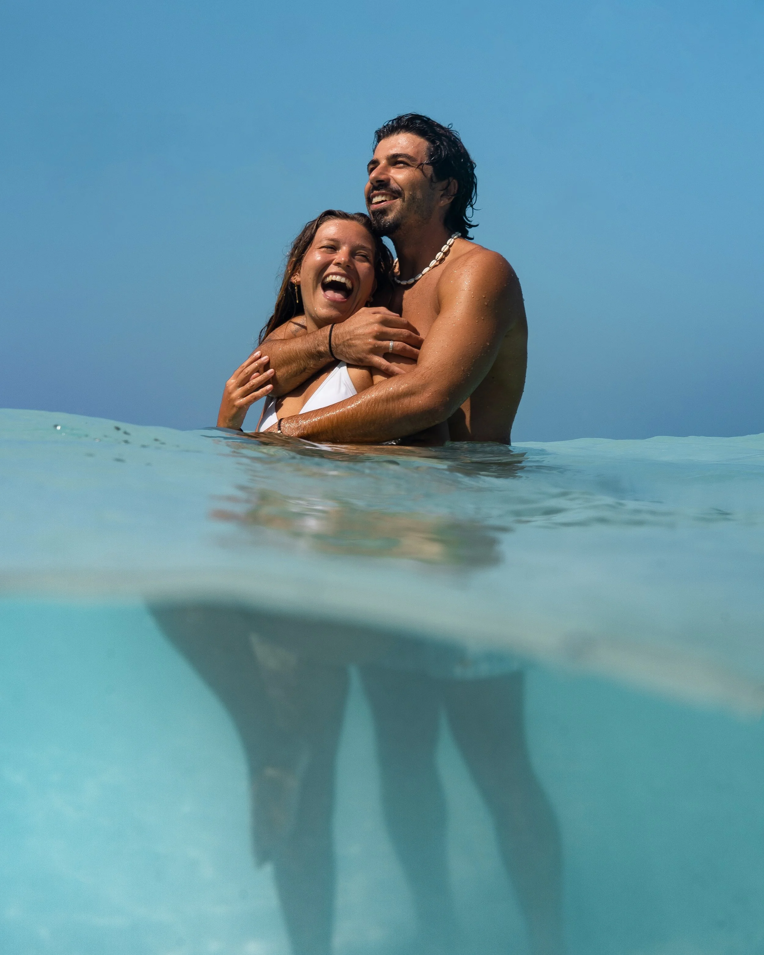 A smiling couple is embracing in shallow ocean water under a clear blue sky. The woman has long brown hair and is wearing a white swimsuit, while the man has dark curly hair, a goatee, and is wearing a shell necklace.