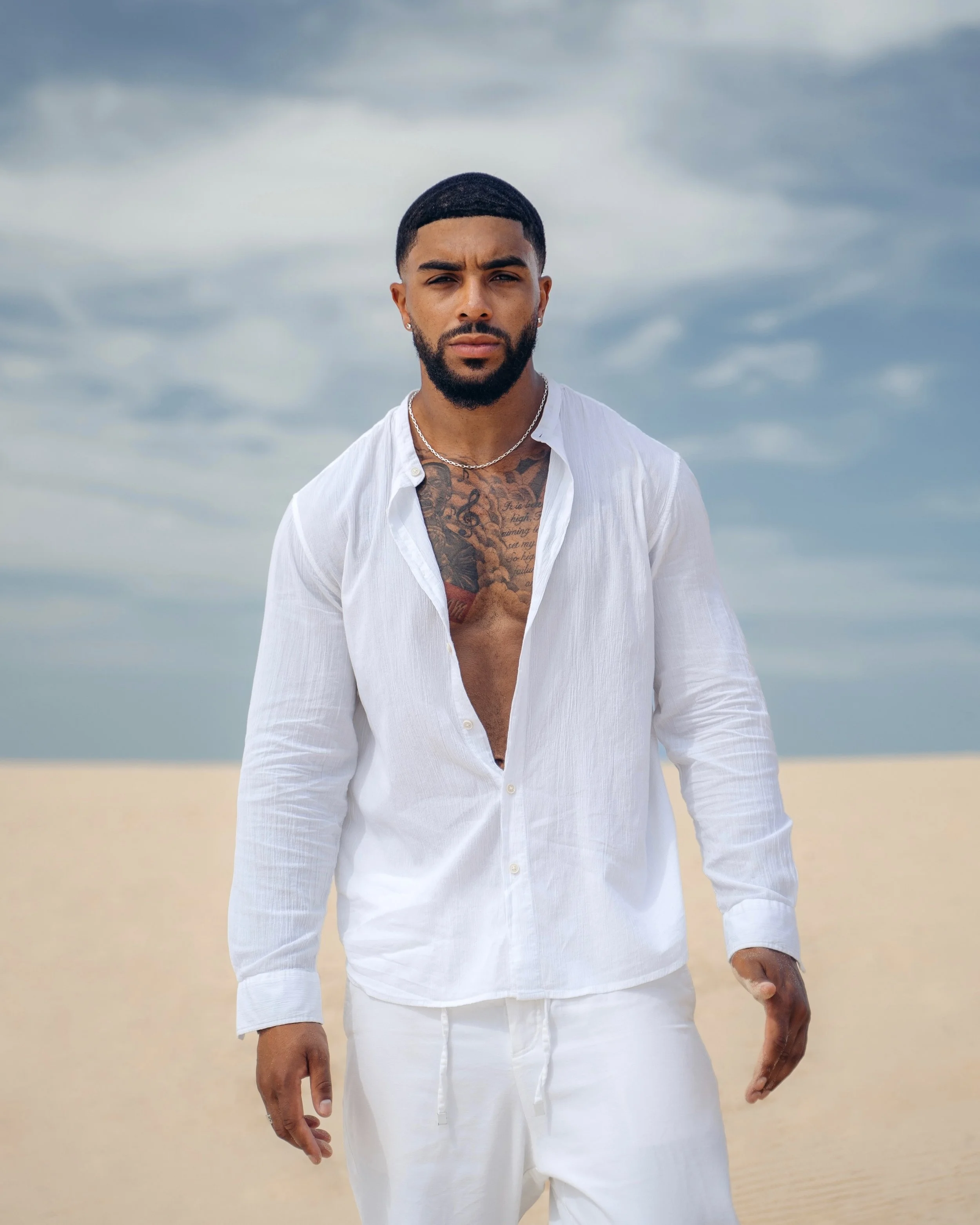 A man with a beard and tattoos on his chest is standing on a beach with cloudy skies in the background. He is wearing a partially unbuttoned white shirt and white pants.