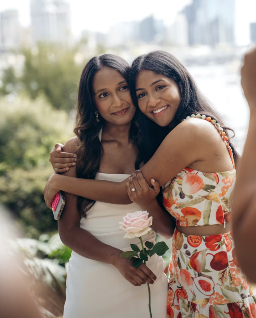 Two women hugging and smiling, one holding a pink rose, in an outdoor setting with a cityscape background.