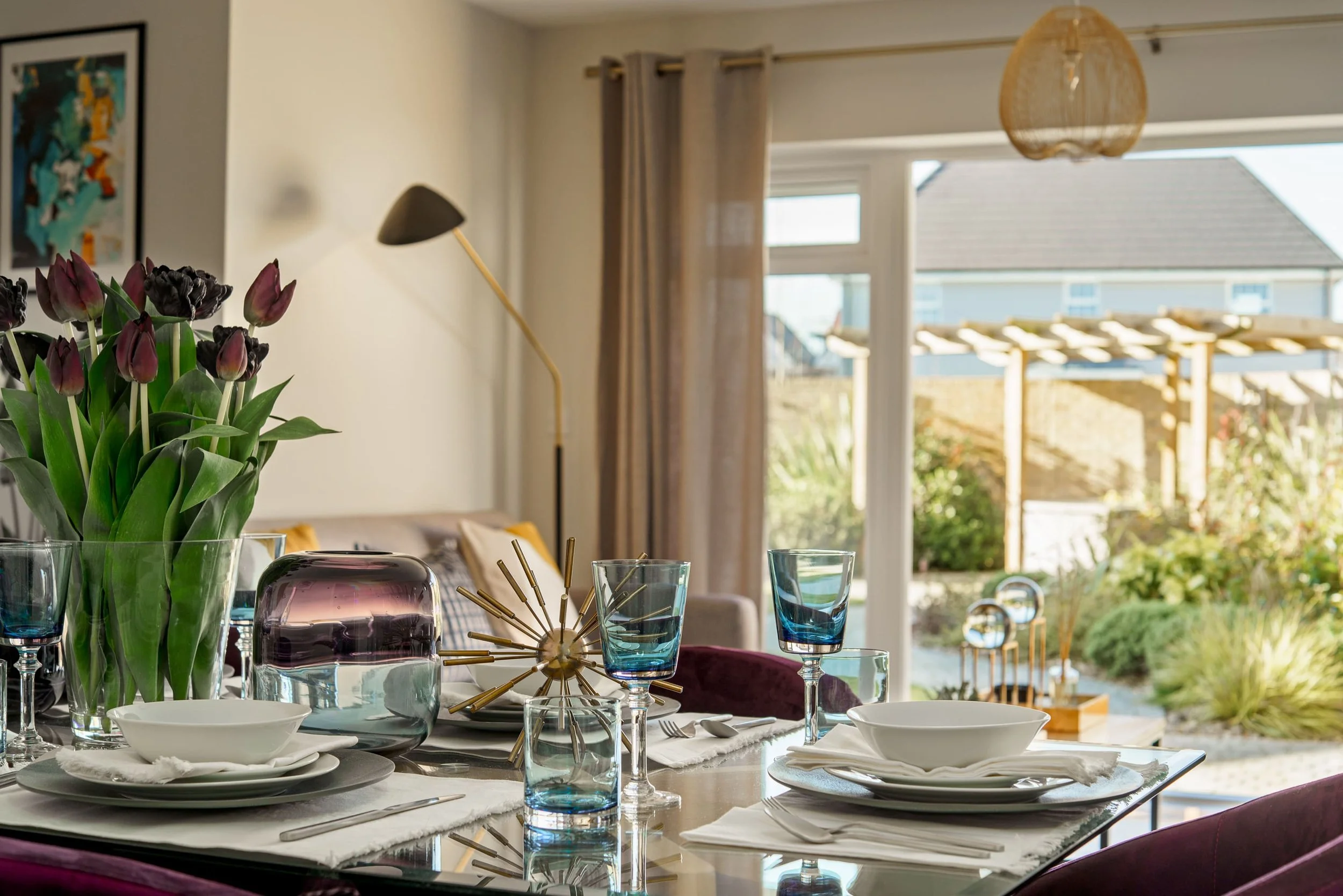 A dining table set with white plates, blue glasses, and a centerpiece with purple tulips in a clear vase. The room has large windows with beige curtains, allowing natural light to fill the space. Some decorative objects and artwork are visible in the background.