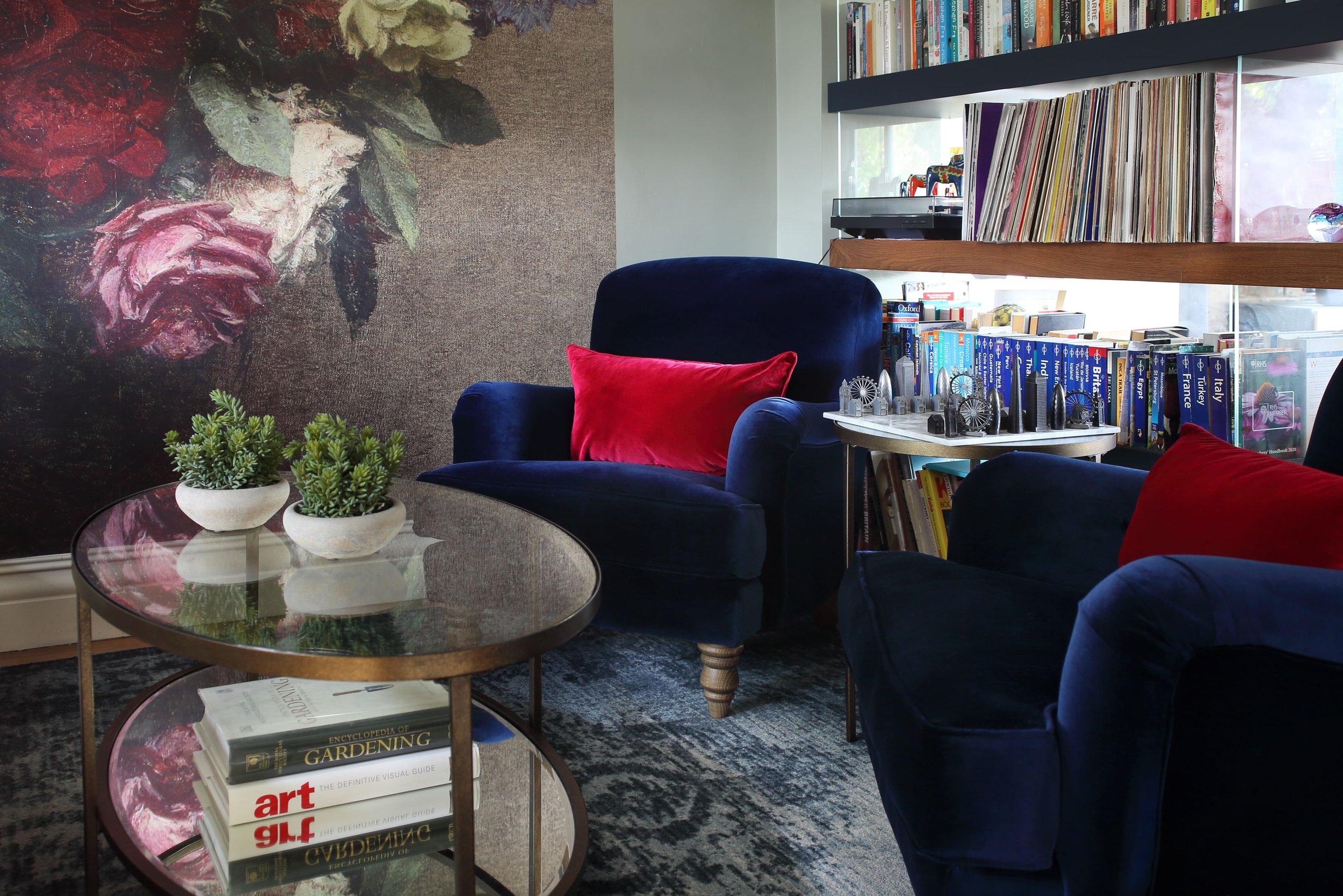 Interior view of a cozy reading corner with a blue velvet armchair with a red pillow, a round glass coffee table with books and small potted plants, a large floral wall art, and a bookshelf filled with books, vinyl records, and model Ferris wheels.
