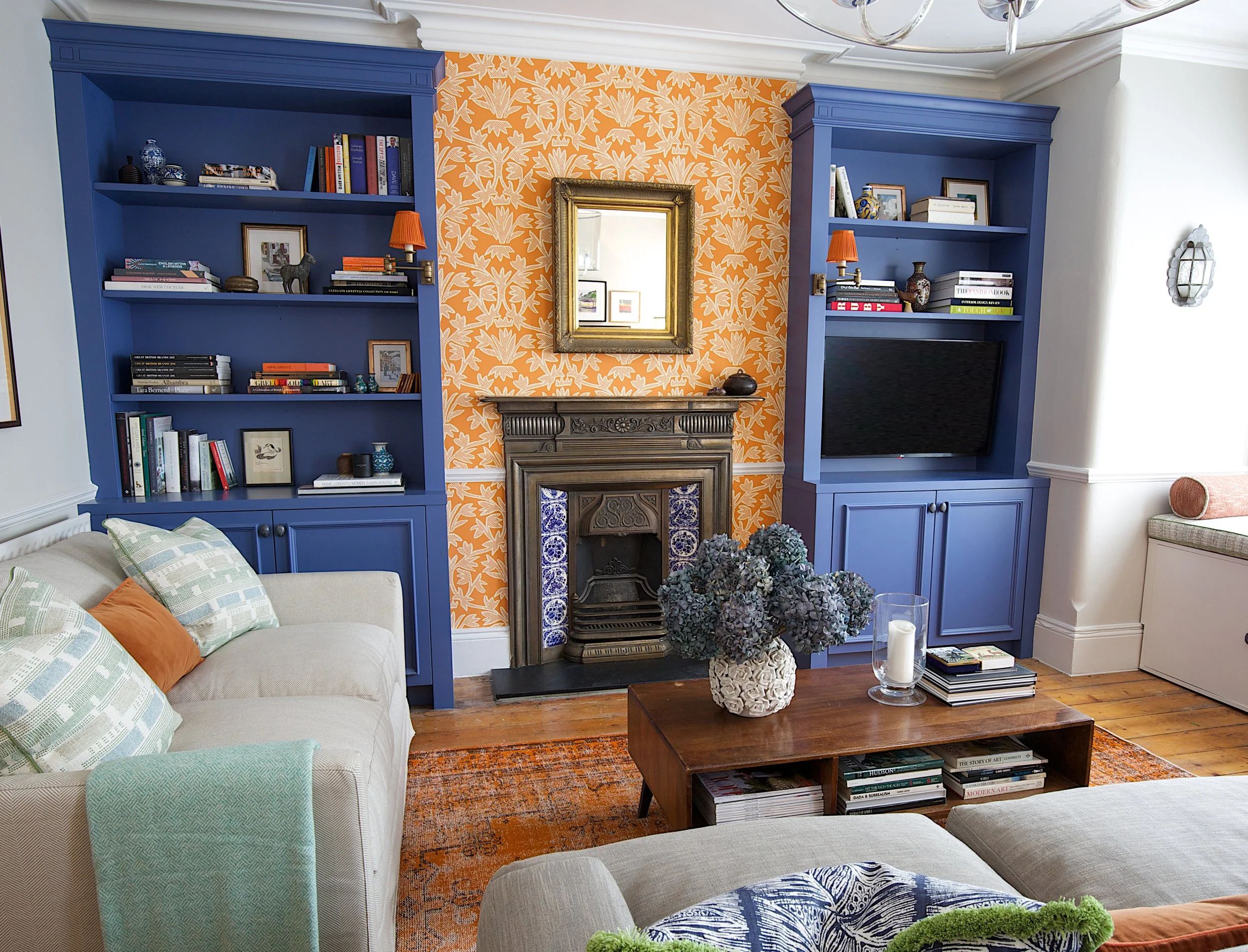 Living room with orange patterned wallpaper, blue built-in bookshelves, a vintage fireplace, mirror, and various decorative items, with a beige sofa and wooden coffee table.