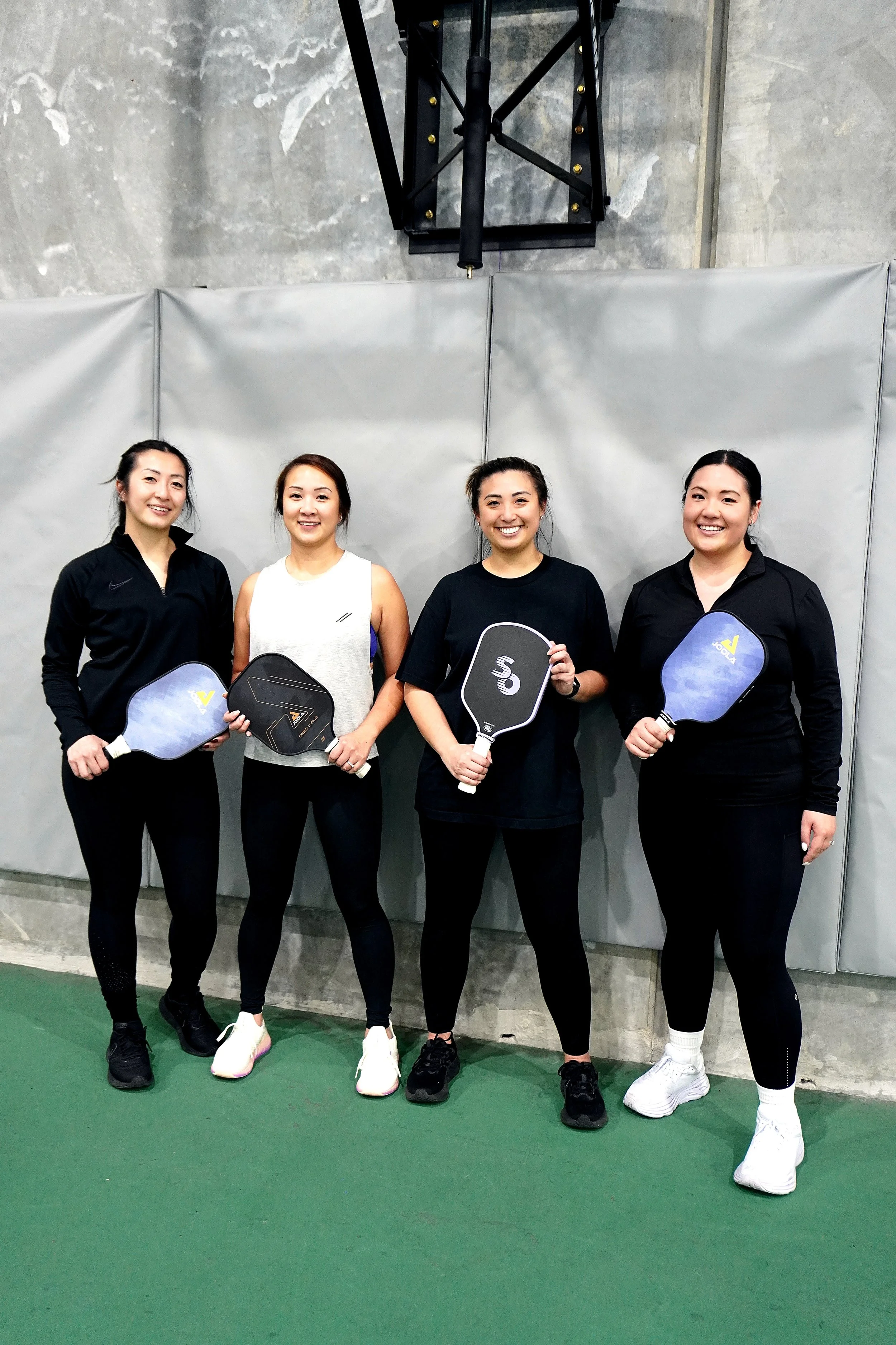 Four women standing side by side indoors holding pickleball paddles, dressed in athletic clothing, against a gray wall with a black structure overhead and green floor.