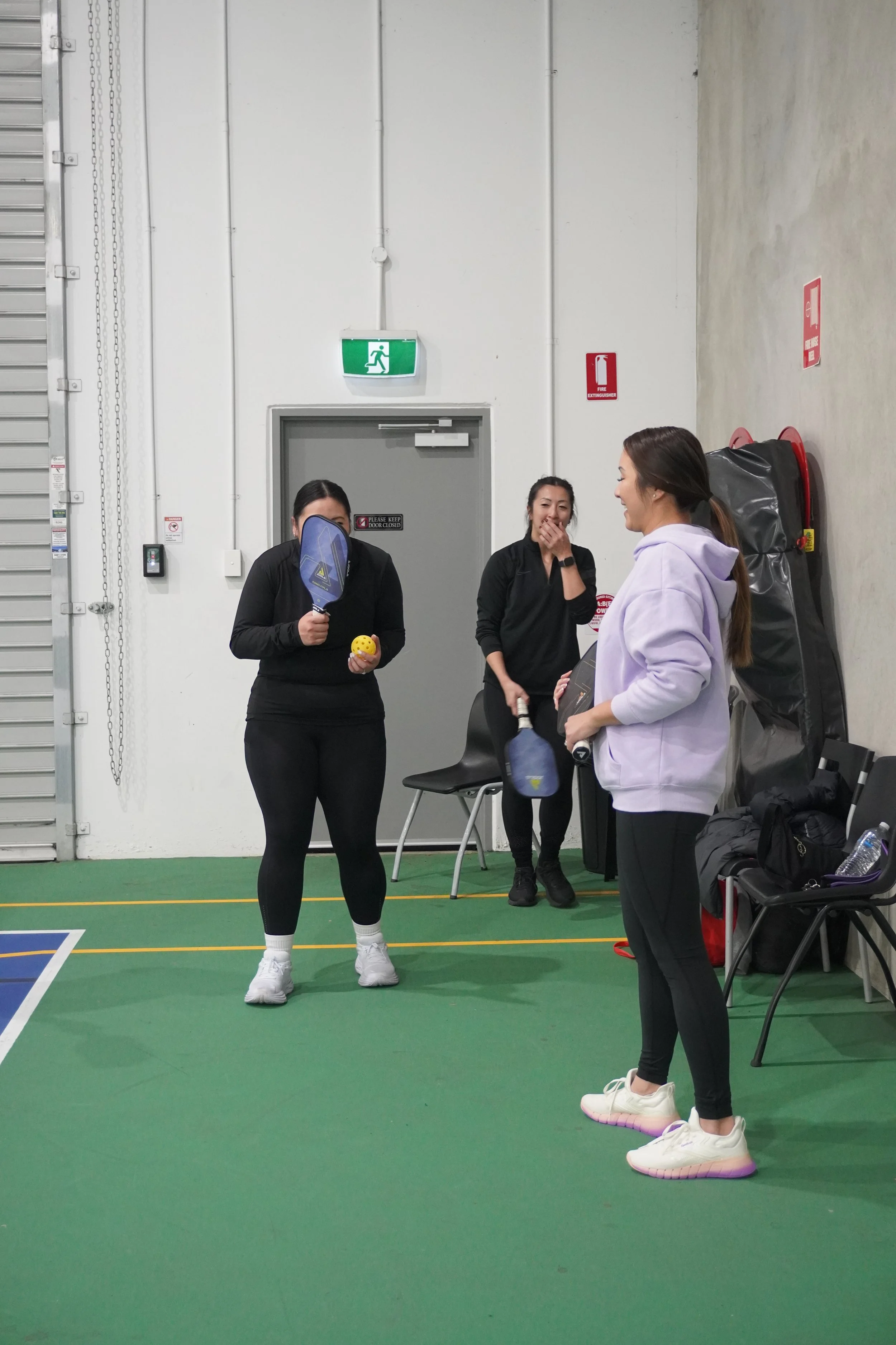 Three women standing and conversing inside an indoor sports facility. One woman holds a ping pong paddle and ball while the other two smile and talk. They are casually dressed, and there are chairs, a fire extinguisher, and emergency exit signs visible.