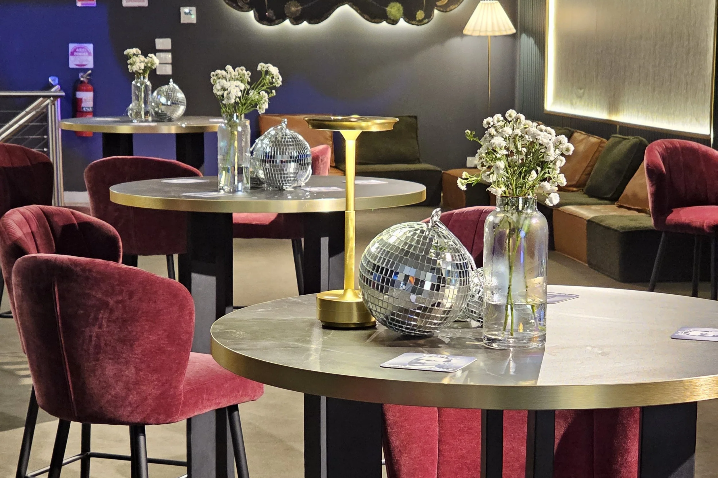 A seating area in a stylish room with a round table decorated with a vase of white flowers and decorative disco balls. Red velvet chairs surround the table, and a long sofa with multicolored cushions is visible in the background. A yellow floor lamp and a dark wall with circular wall art add to the chic ambiance.