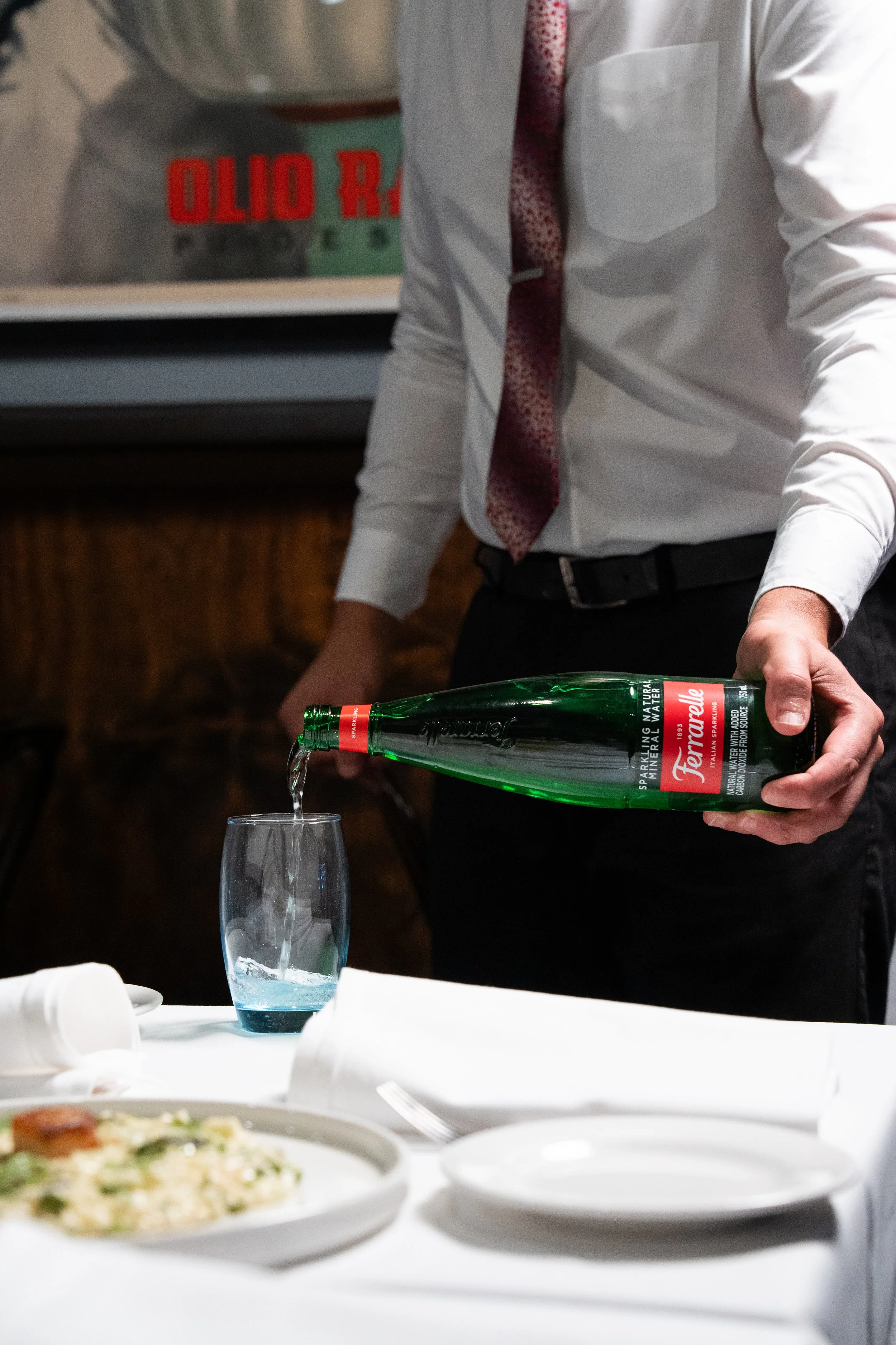 Person in a white shirt and red patterned tie pouring sparkling water into a glass at a dining table with dishes and plates.