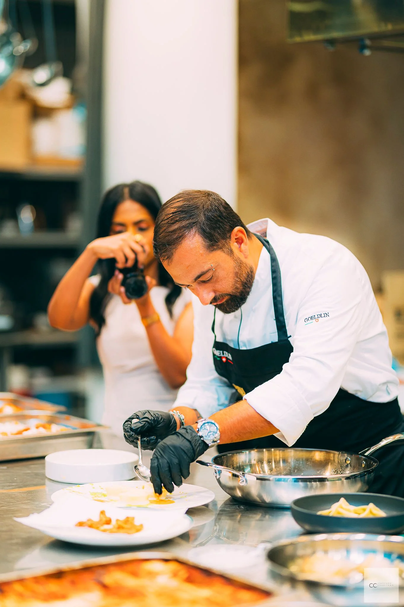 A male chef with dark hair and beard wearing black gloves, a white chef's coat with