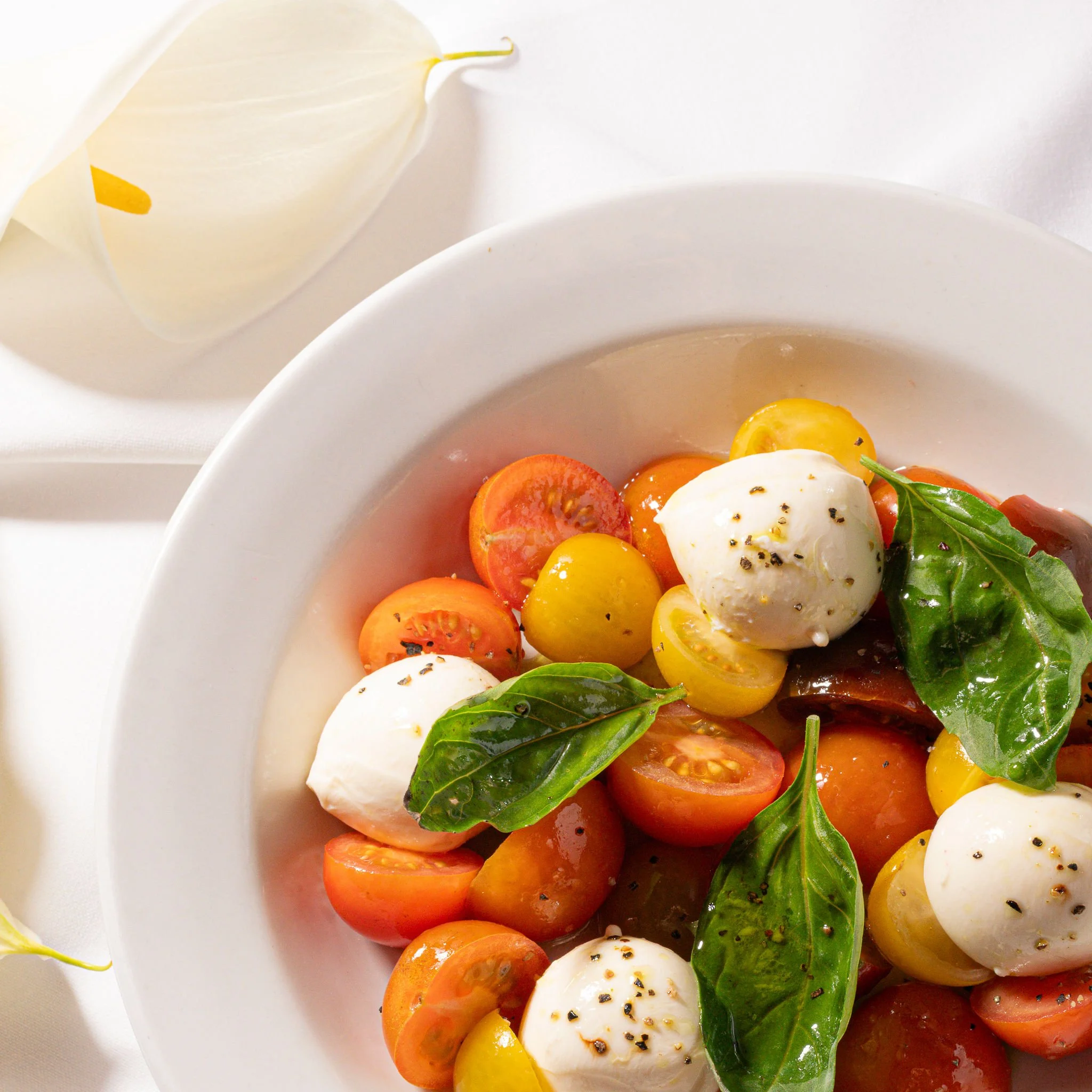 A plate of sliced cherry and grape tomatoes, fresh basil leaves, and mozzarella balls, seasoned with black pepper, on a white plate.