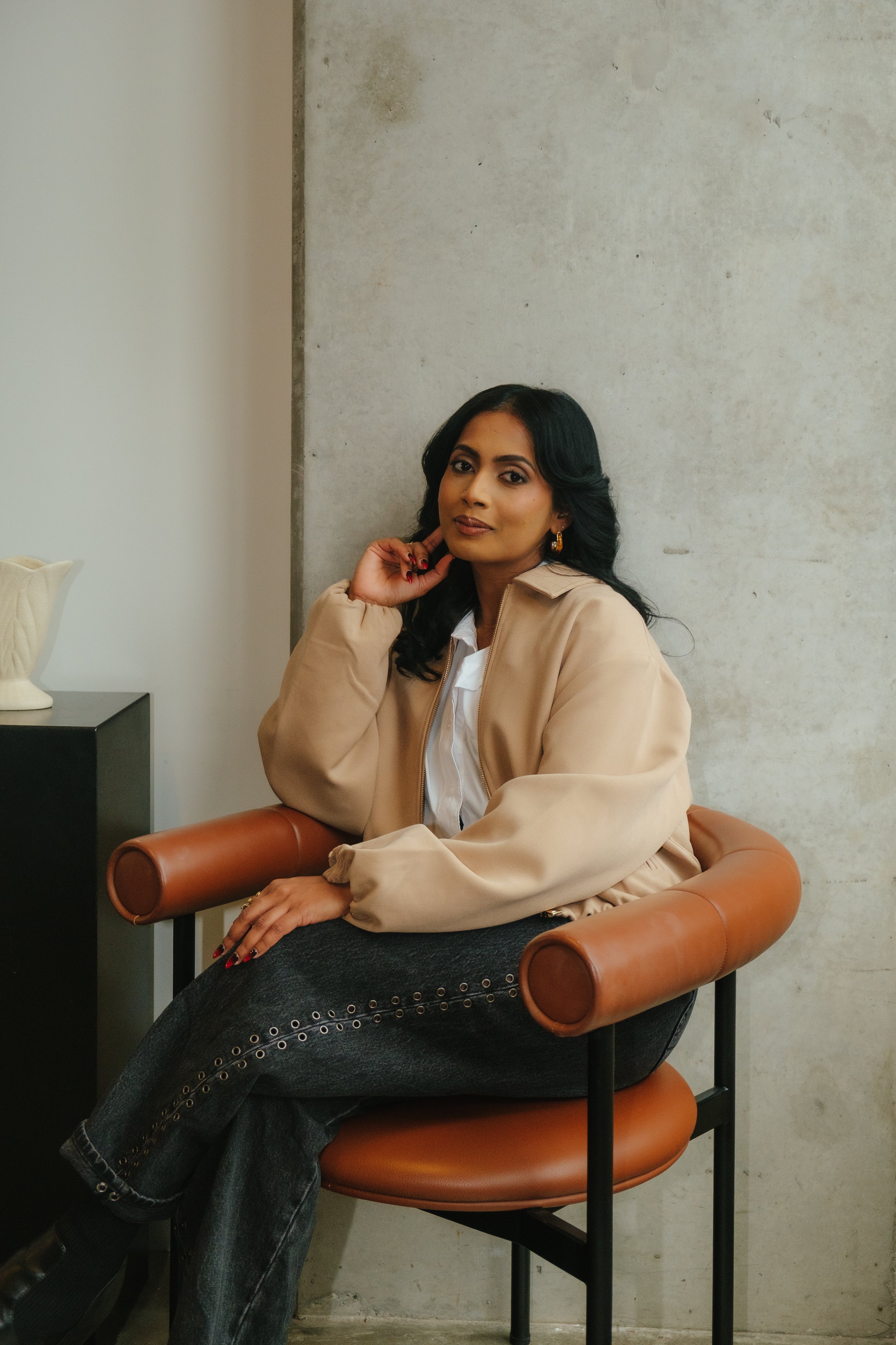 A woman with dark hair sitting on a tan armchair, wearing a light beige jacket, white shirt, and dark jeans, with a neutral expression, posing in front of a plain, textured wall.