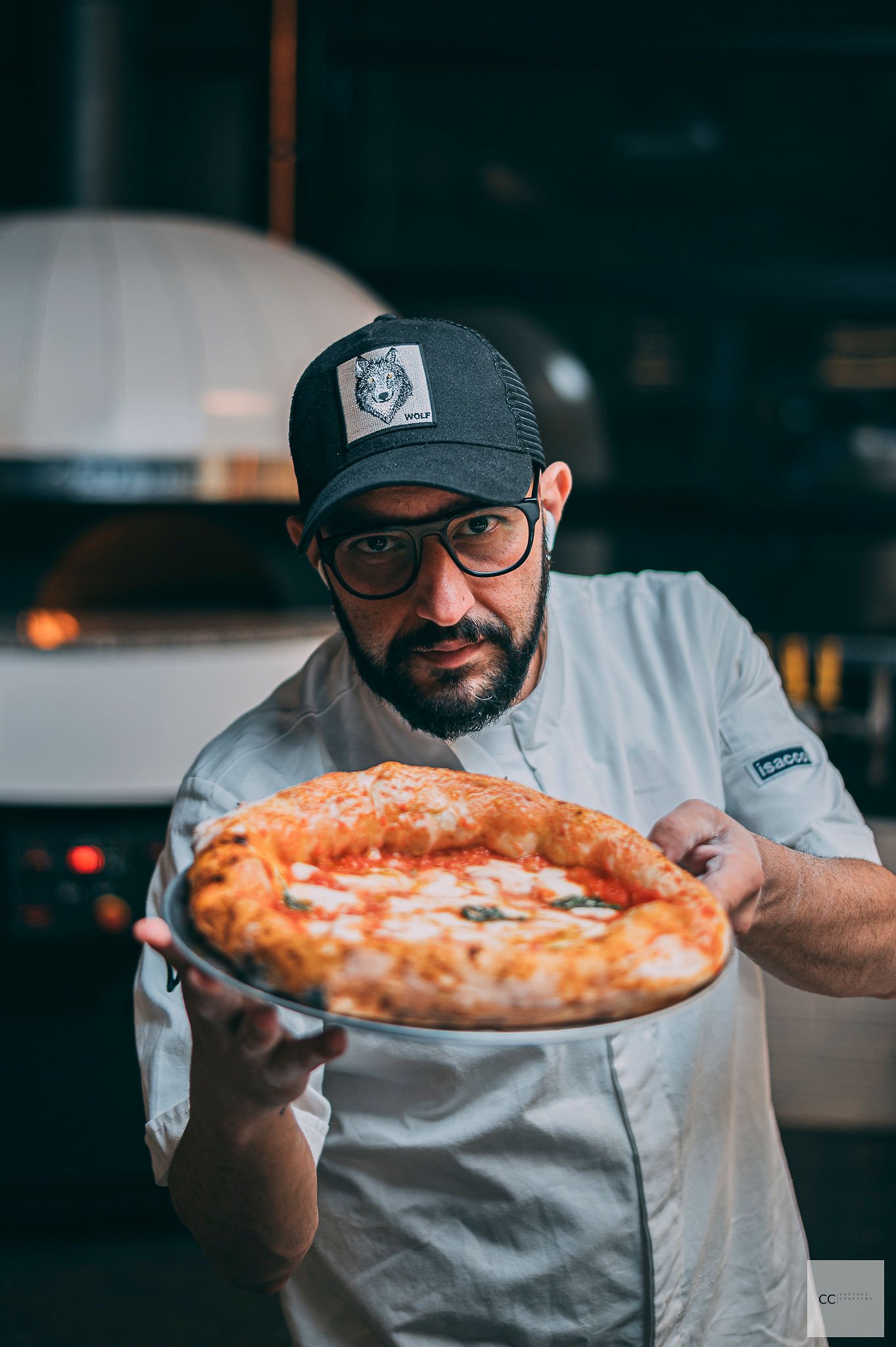 A chef with glasses and a beard wearing a black cap with a wolf patch, holding a pizza with cheese and basil, in a restaurant kitchen.
