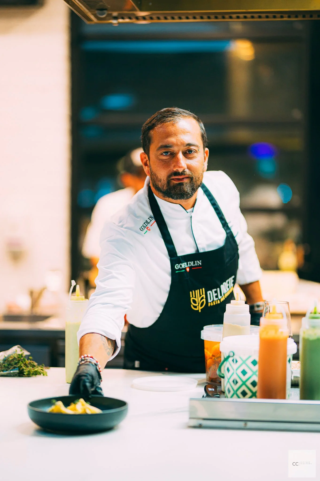 A male chef with a beard and short hair working in a professional kitchen, wearing a white chef coat and black apron that has 'Greatest Chef' printed on it, surrounded by condiment bottles and food ingredients.