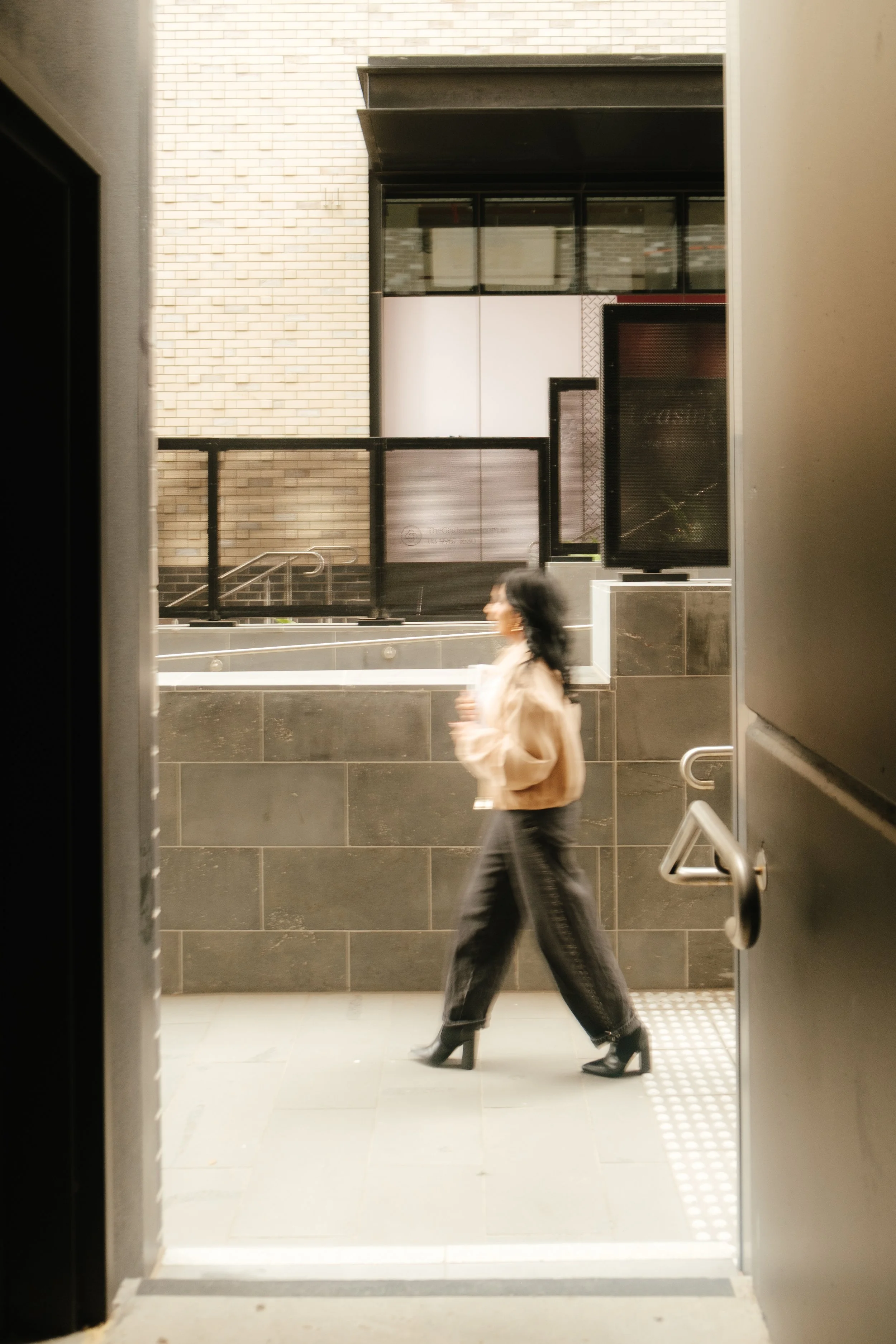 A woman walking through an indoor area, seen from an open door frame. She is wearing a beige jacket, black pants, and black heeled boots. The background includes a brick wall, black railing, and a TV screen.