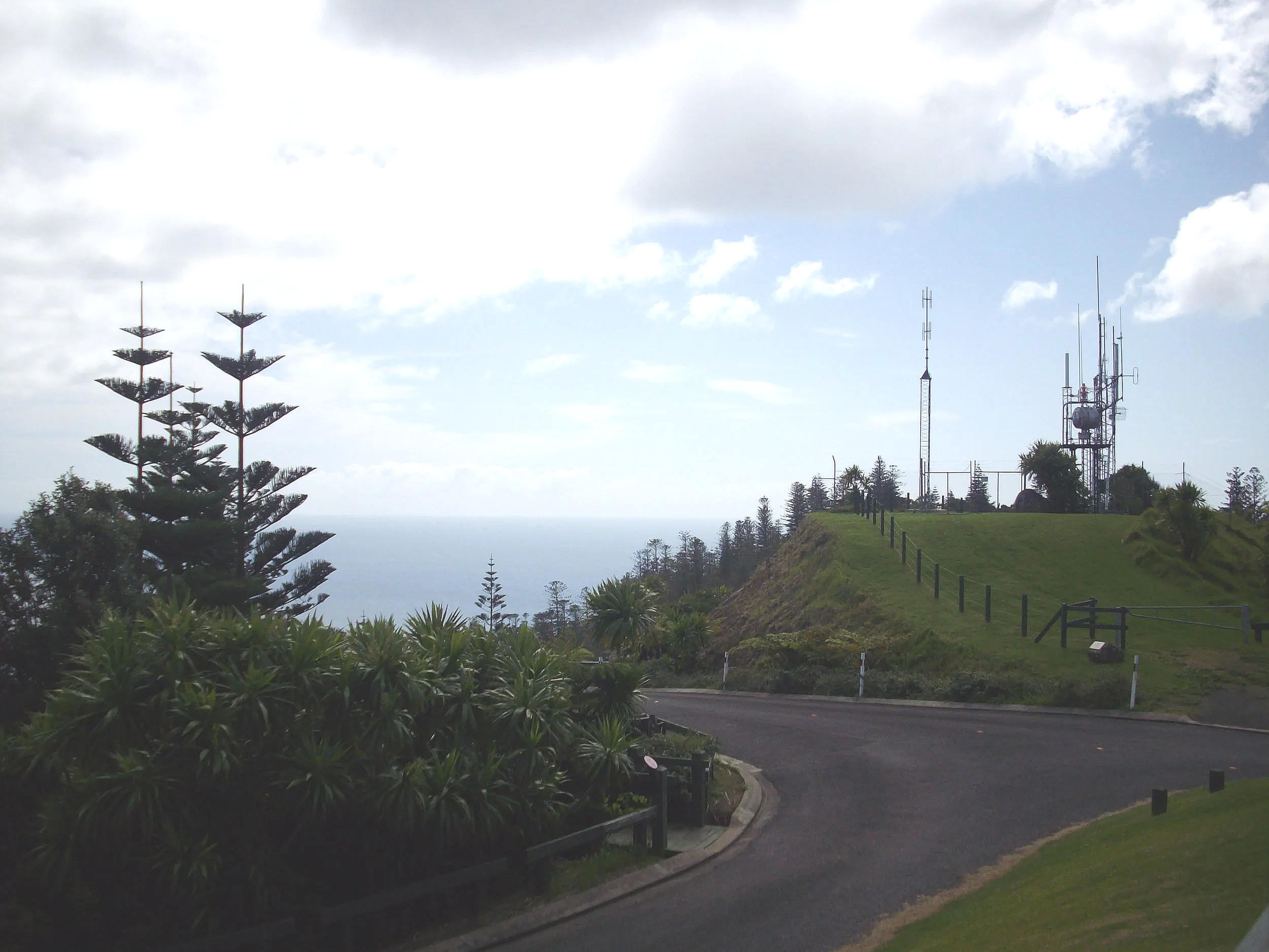 A hilly landscape with tall communication towers, trees, and a cloudy sky.