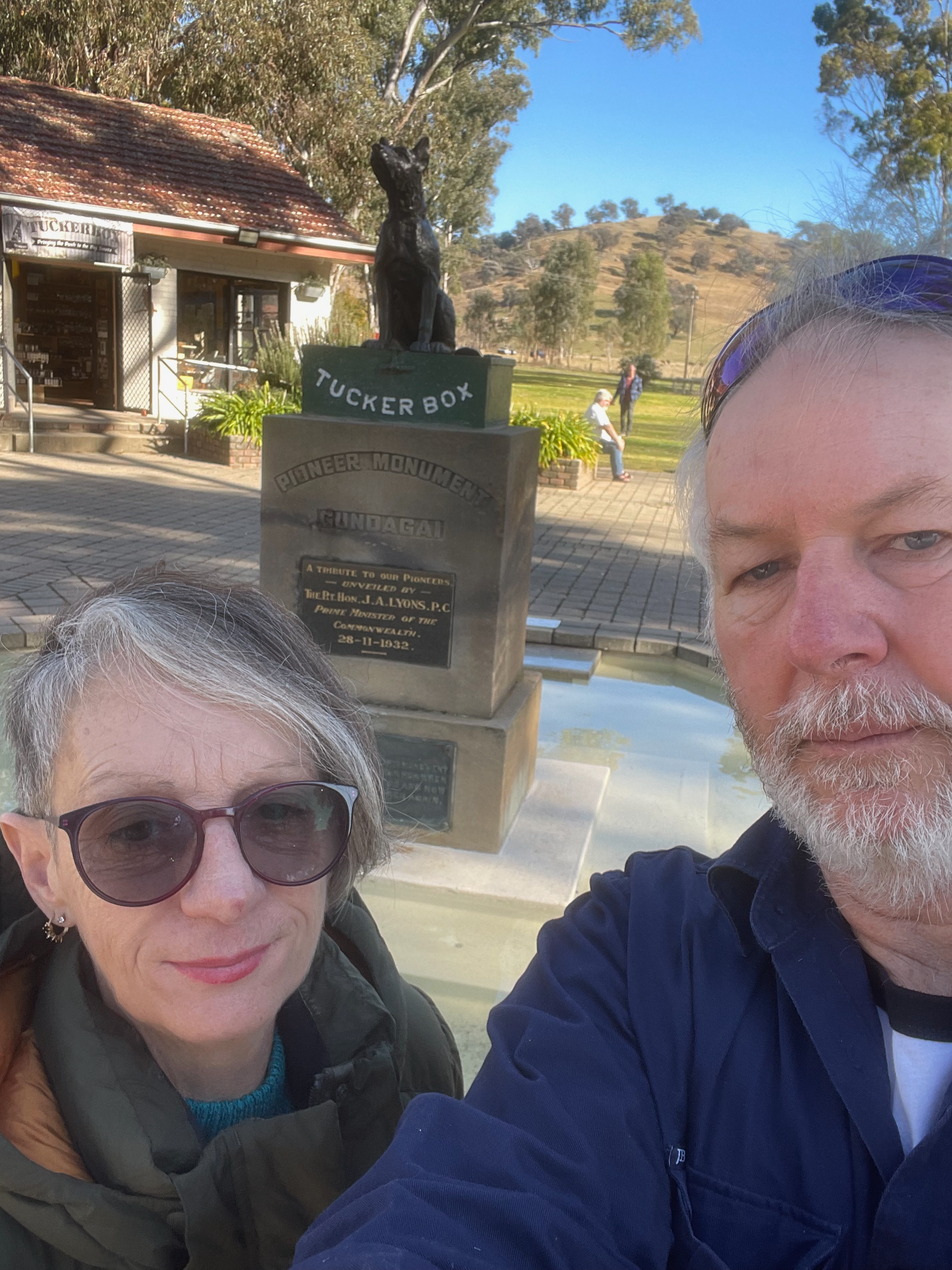 A man and woman taking a selfie outdoors near a monument with a statue of a dog and a plaque, with trees and a hill in the background.