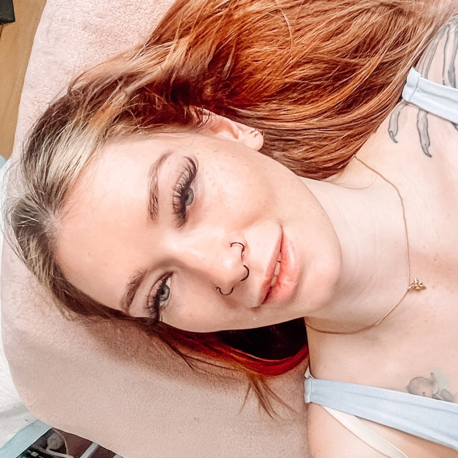 A woman with red hair lying on a bed with her eyes closed, wearing a septum piercing, a gold necklace, and a white tank top.