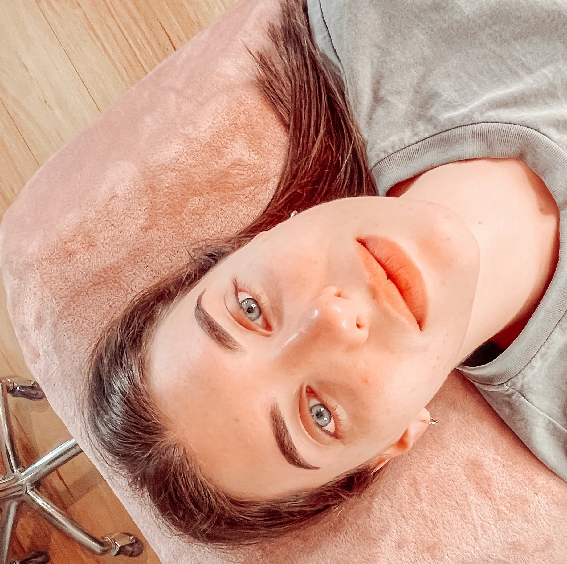 A person with light skin lying on a towel with their eyes open, with their head resting on a folded towel, near some wooden furniture and chrome ladders.