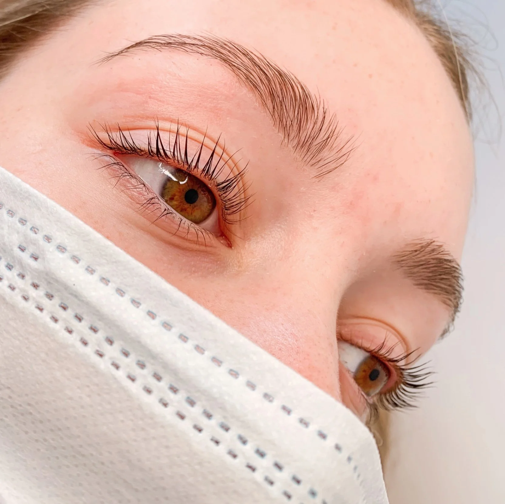 Close-up of a person's face with brown eyes, long eyelashes, well-groomed eyebrows, and light skin, partially covered by a white medical face mask.