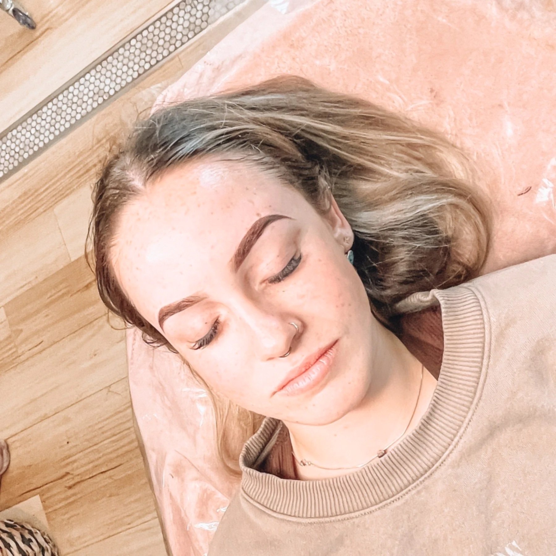 Close-up of a woman's face lying on a pink marble surface with her eyes closed and hair spread out, wearing a beige sweatshirt.