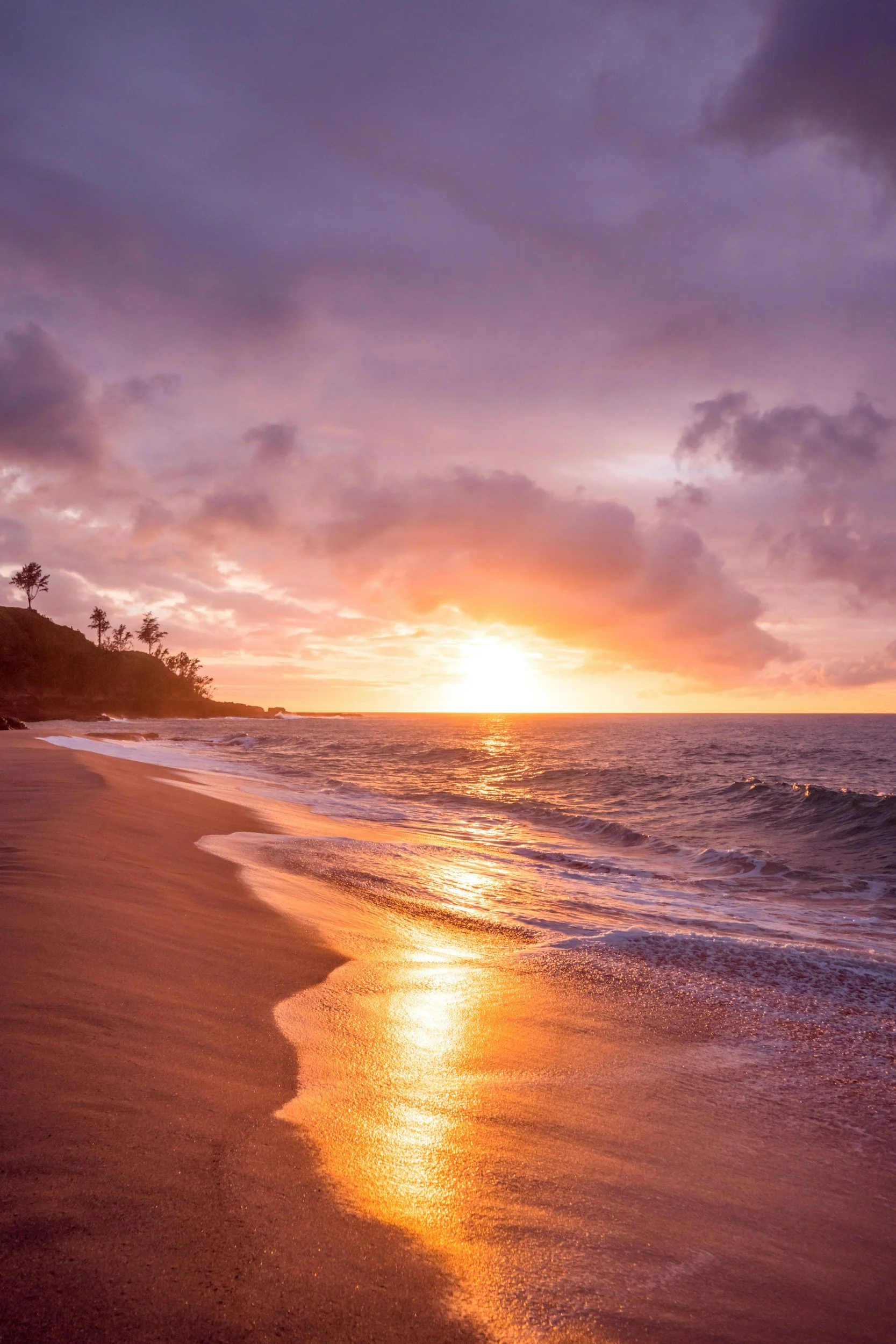 Sunset over a beach with purple and pink clouds in the sky, reflecting orange light on the wet sand, with small waves rolling onto the shore and silhouettes of trees on a hillside.