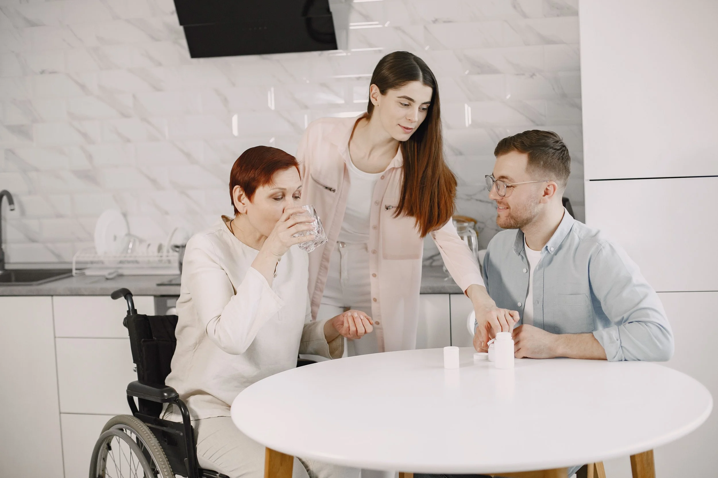 A woman in a wheelchair drinking water at a white table with a man and a caregiver in a kitchen.
