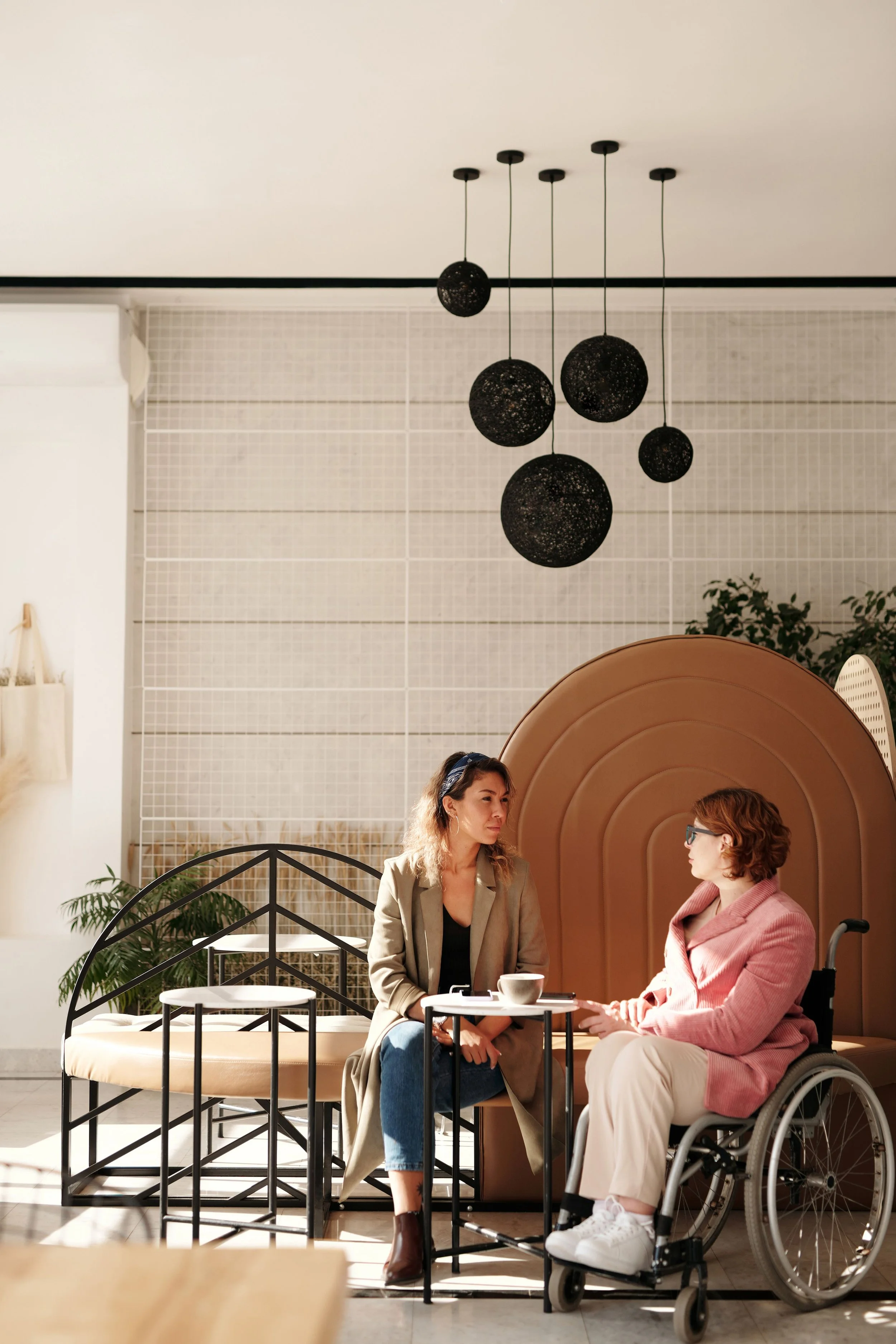Two women sit at a table in a bright room, conversing. One woman is in a wheelchair, and there is a sofa, plants, and modern spherical pendant lights hanging from the ceiling.