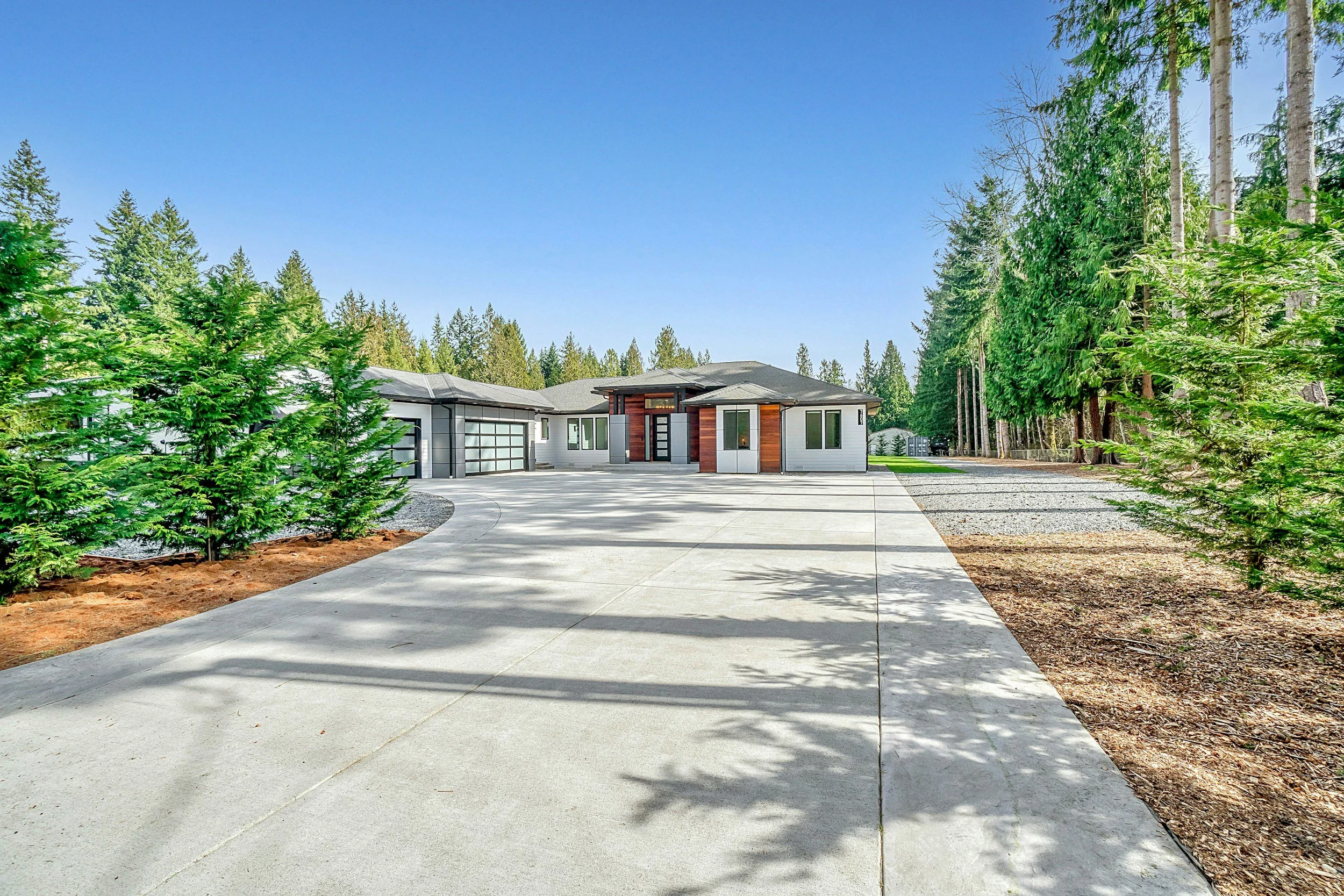 Modern house with a gray roof and white exterior, large glass garage door, surrounded by tall green trees and a concrete driveway on a sunny day.