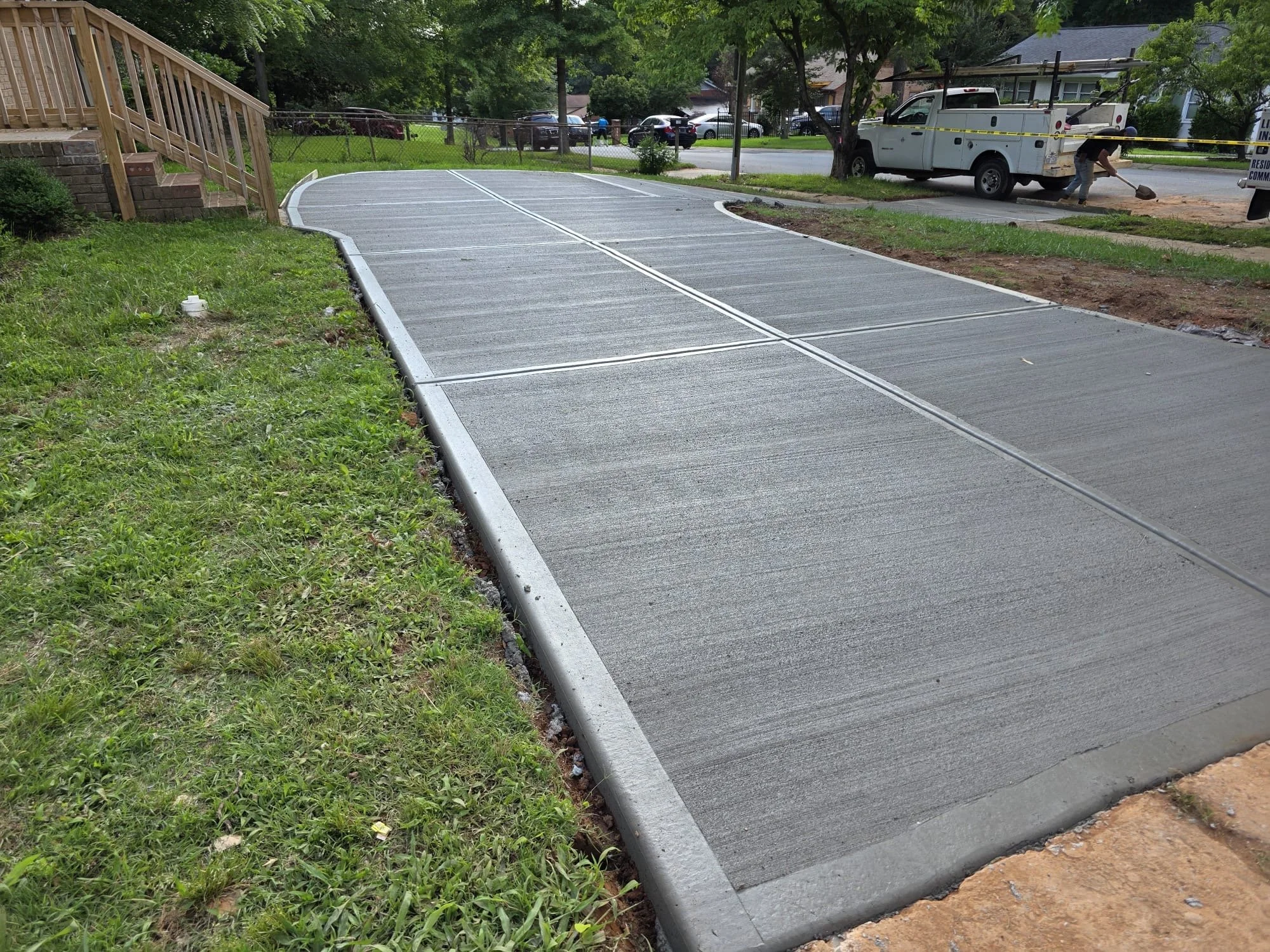 A freshly poured concrete sidewalk with a slight curve leading from a grassy yard to the street, bordered on one side by a wooden staircase and on the other side by dirt and grass. In the background, there is a utility truck and a man working on the sidewalk, with several cars parked along the street and trees providing shade.