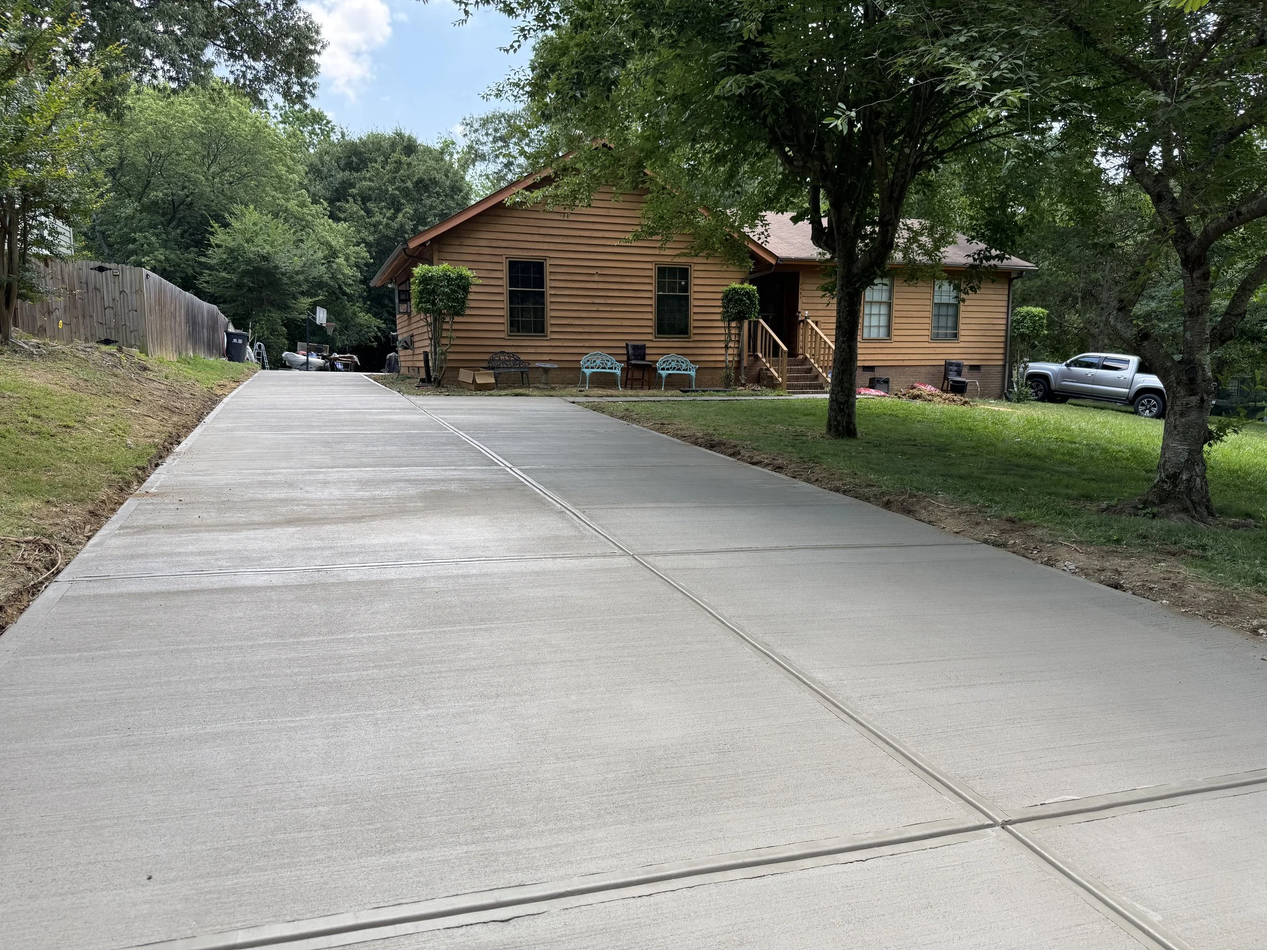 View of a house surrounded by trees, with a driveway leading up to it. There are outdoor chairs and furniture near the house and a pickup truck parked on the grass.