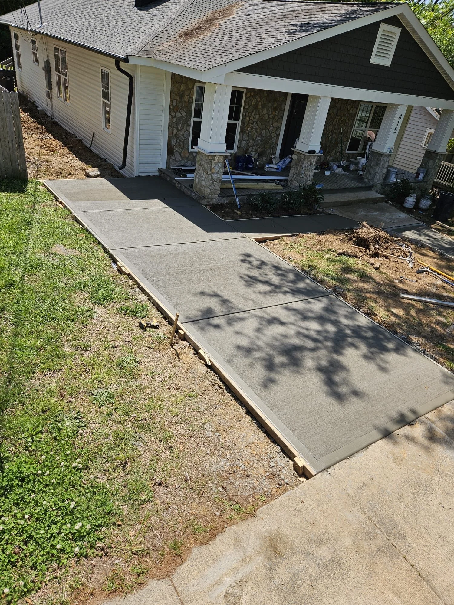 House under construction with freshly poured concrete sidewalk in front, construction tools and materials visible, and landscaping in progress.
