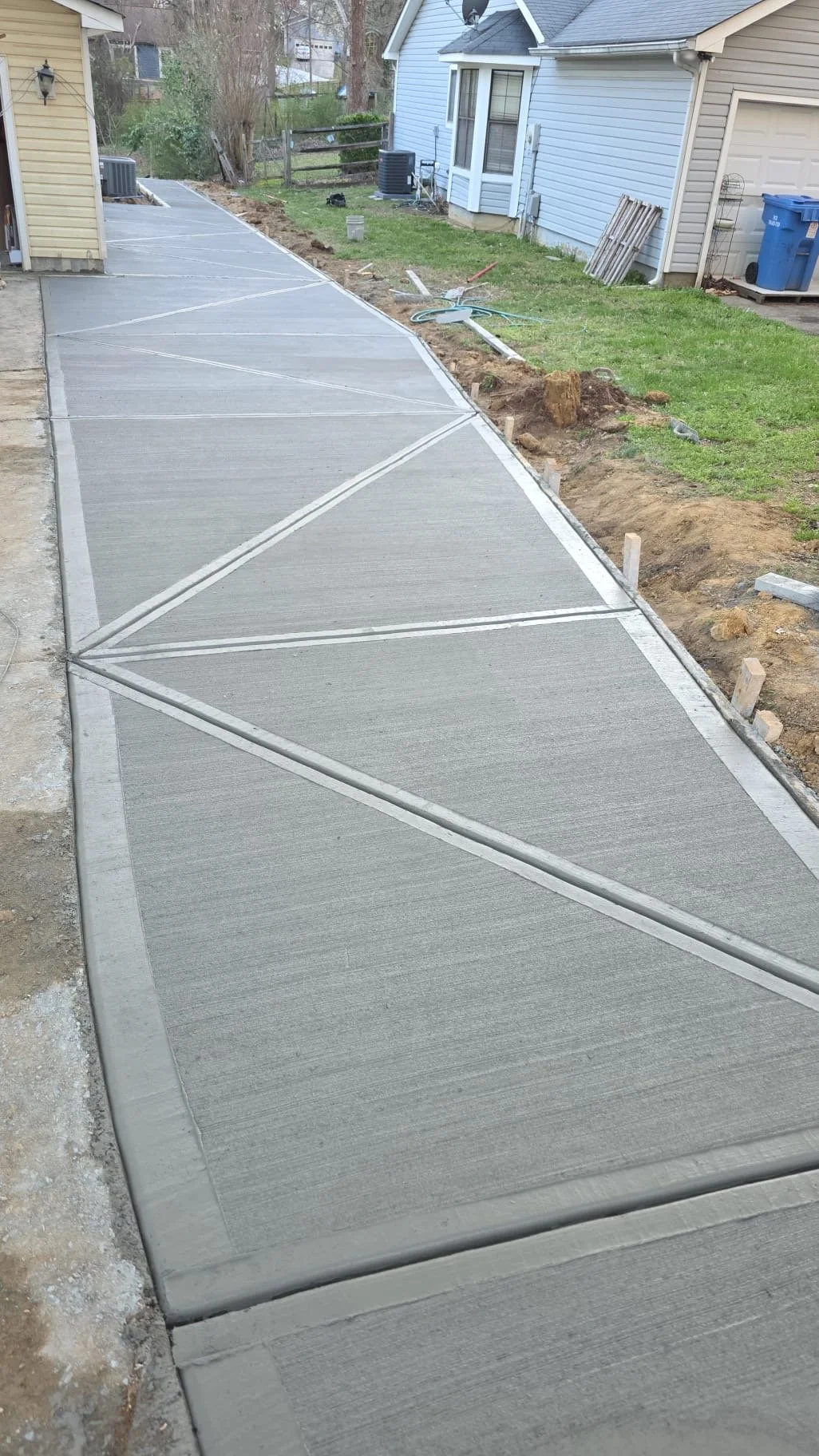 Newly poured concrete sidewalk with edges visible, next to a grass yard and residential houses, some construction tools and materials on the ground.