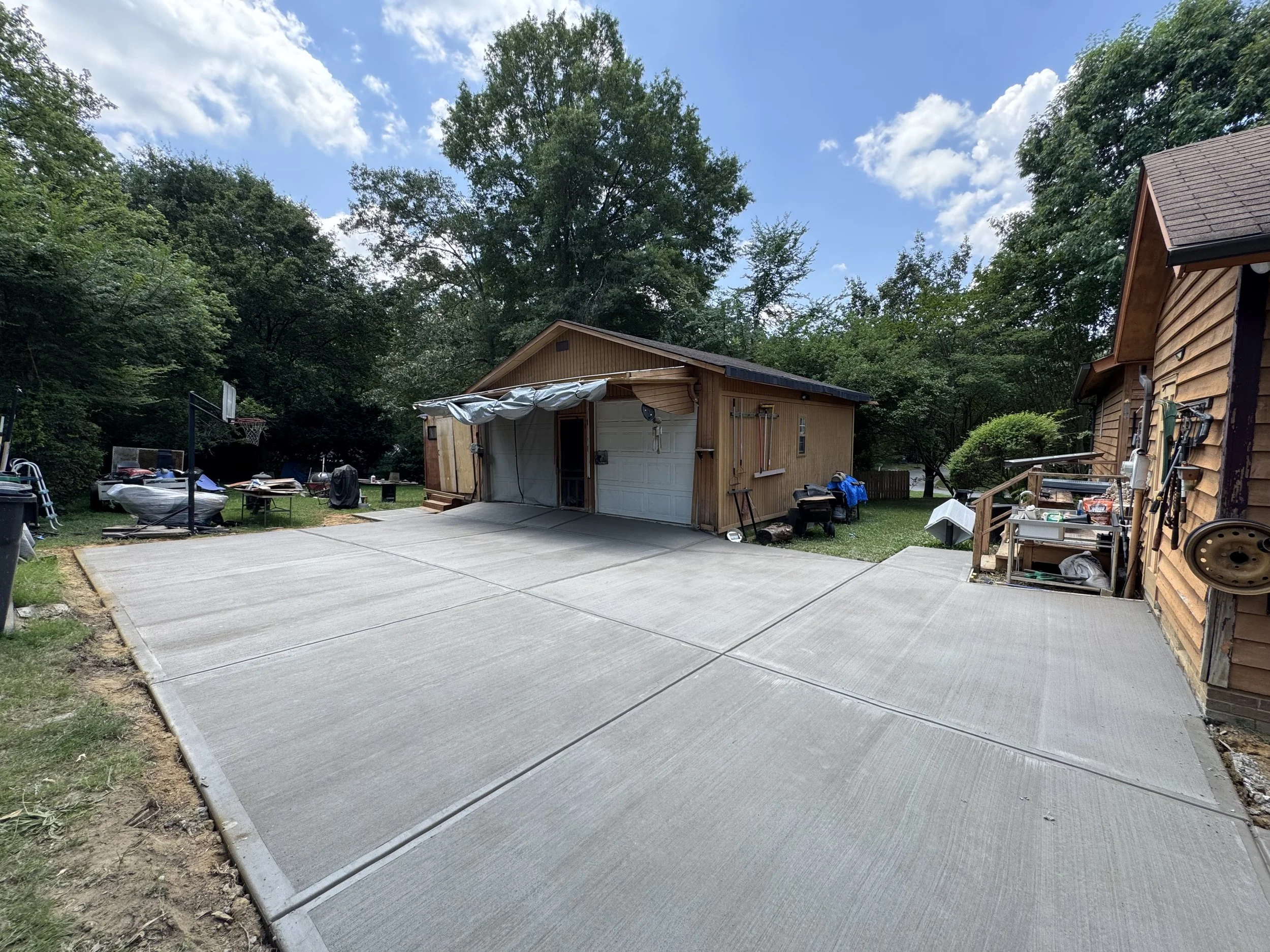 A backyard with a freshly poured concrete driveway and a detached garage surrounded by trees and other buildings.