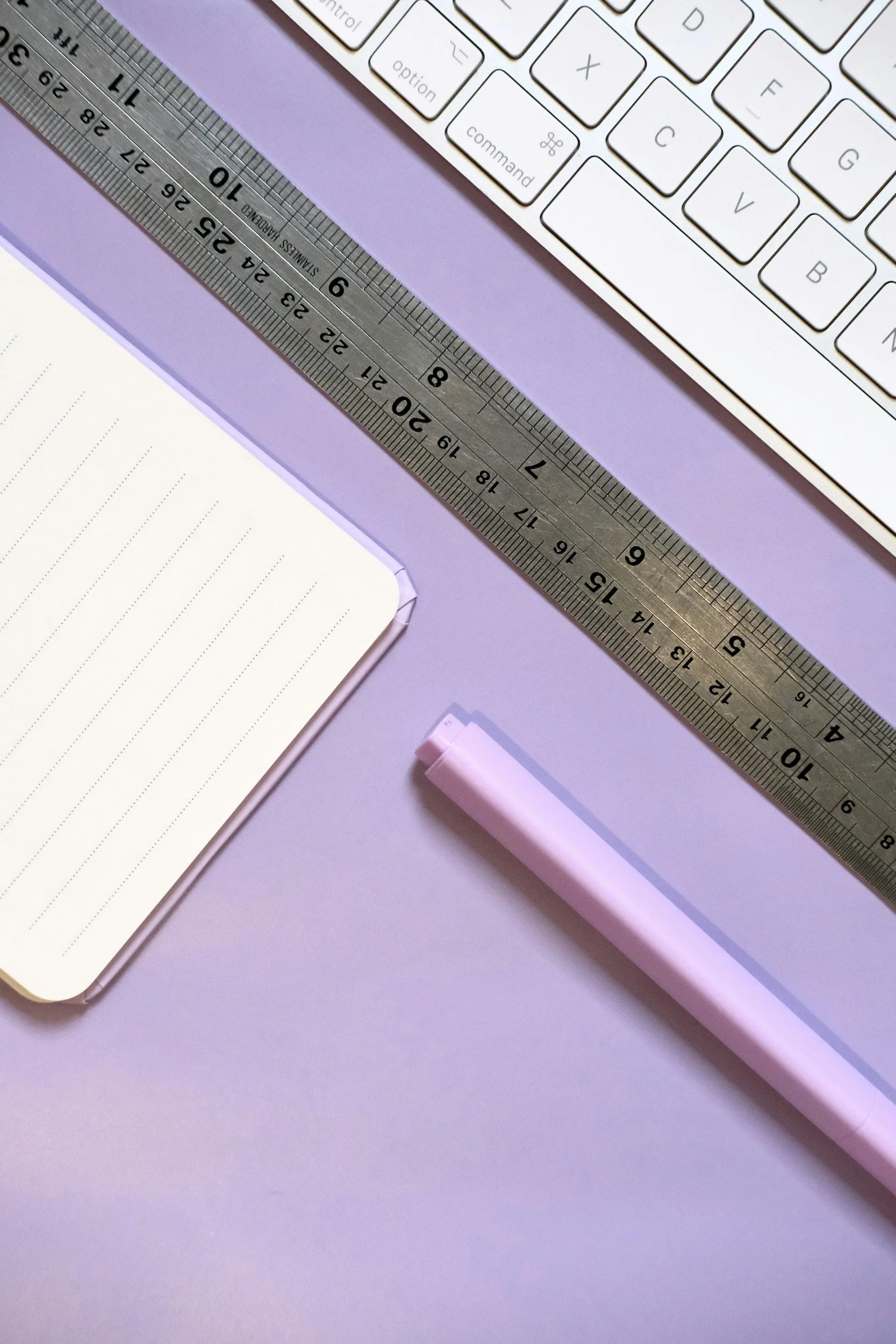 A computer keyboard, a metal ruler, an open notebook with lined pages, and a pink highlighter on a purple desk.