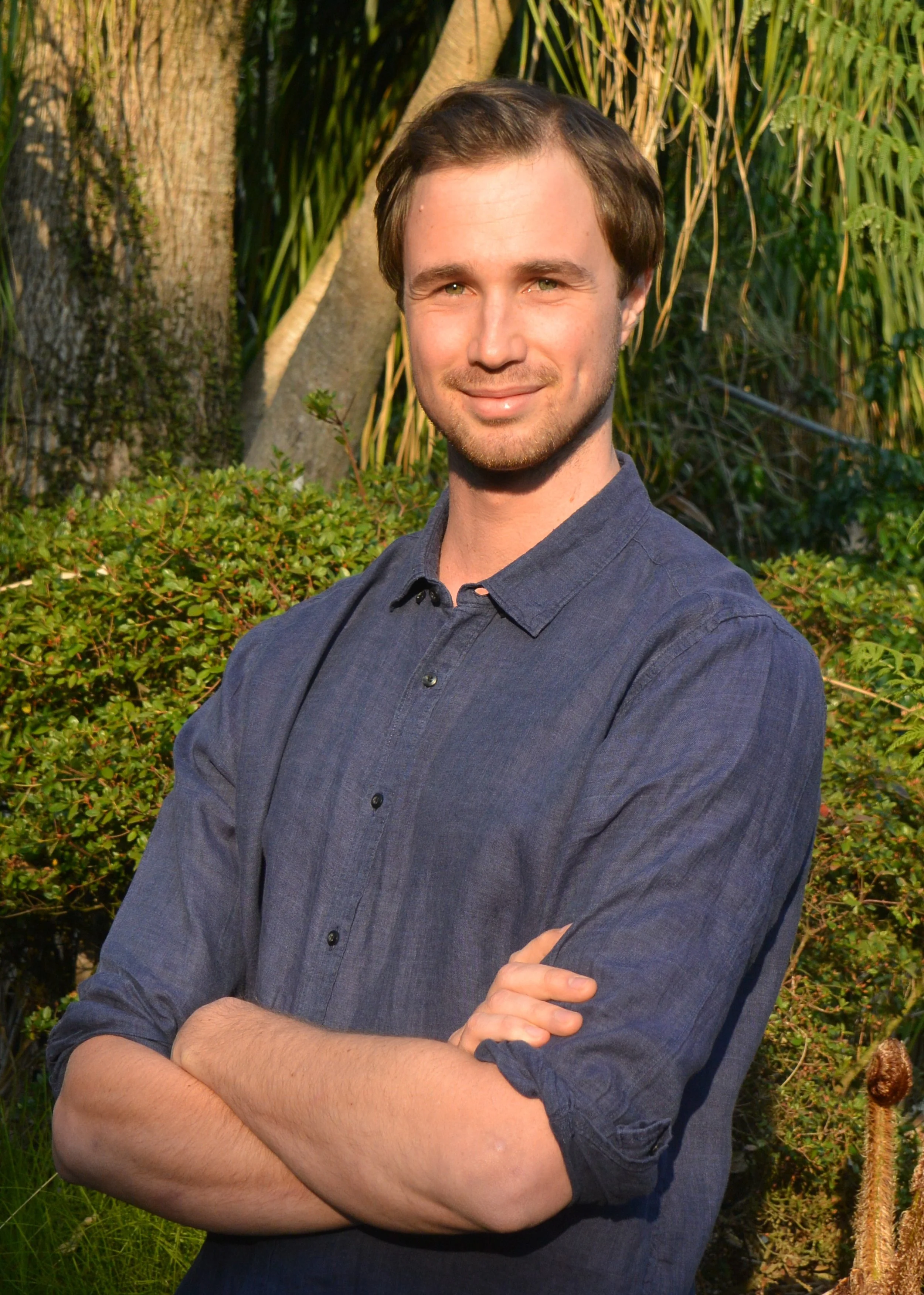 A young man with light skin, brown hair, and green eyes, smiling with arms crossed, standing outdoors in front of trees and bushes.