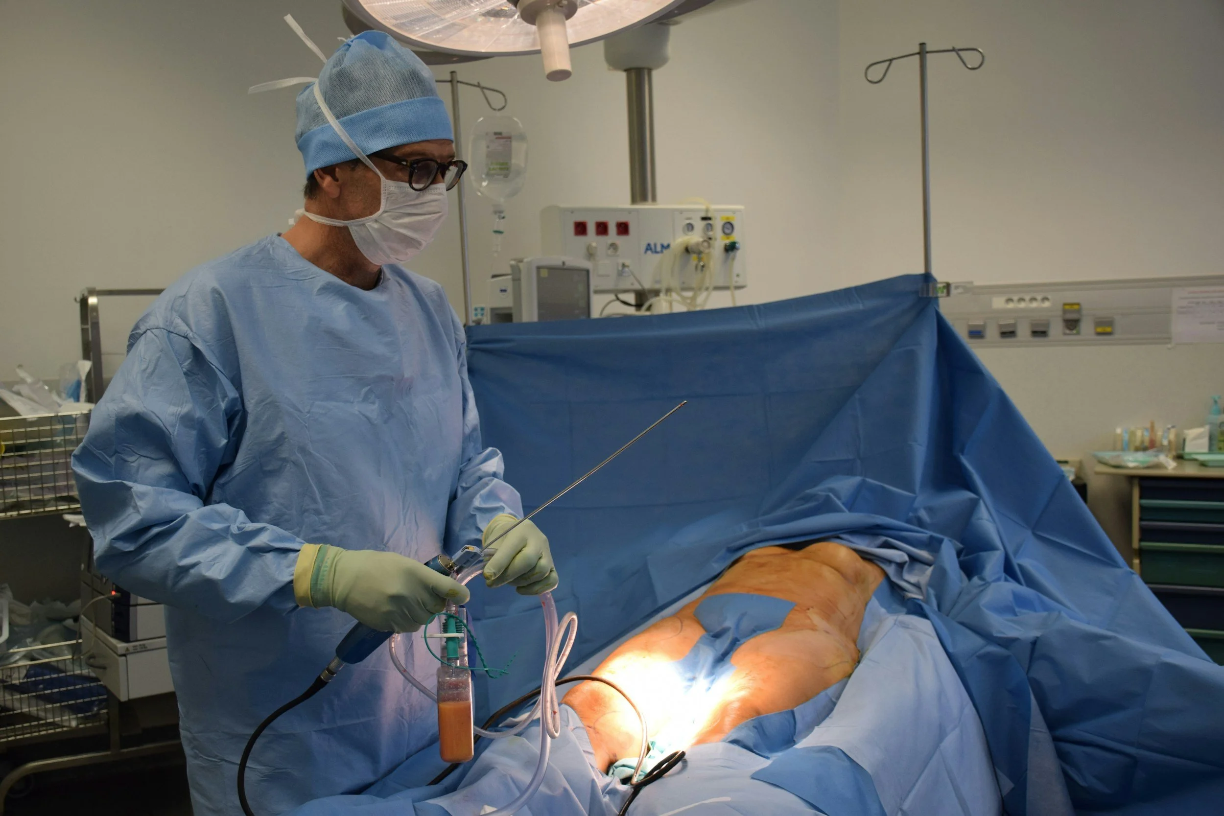 A surgeon in scrubs, mask, and cap performing surgery on a patient in an operating room with medical equipment.
