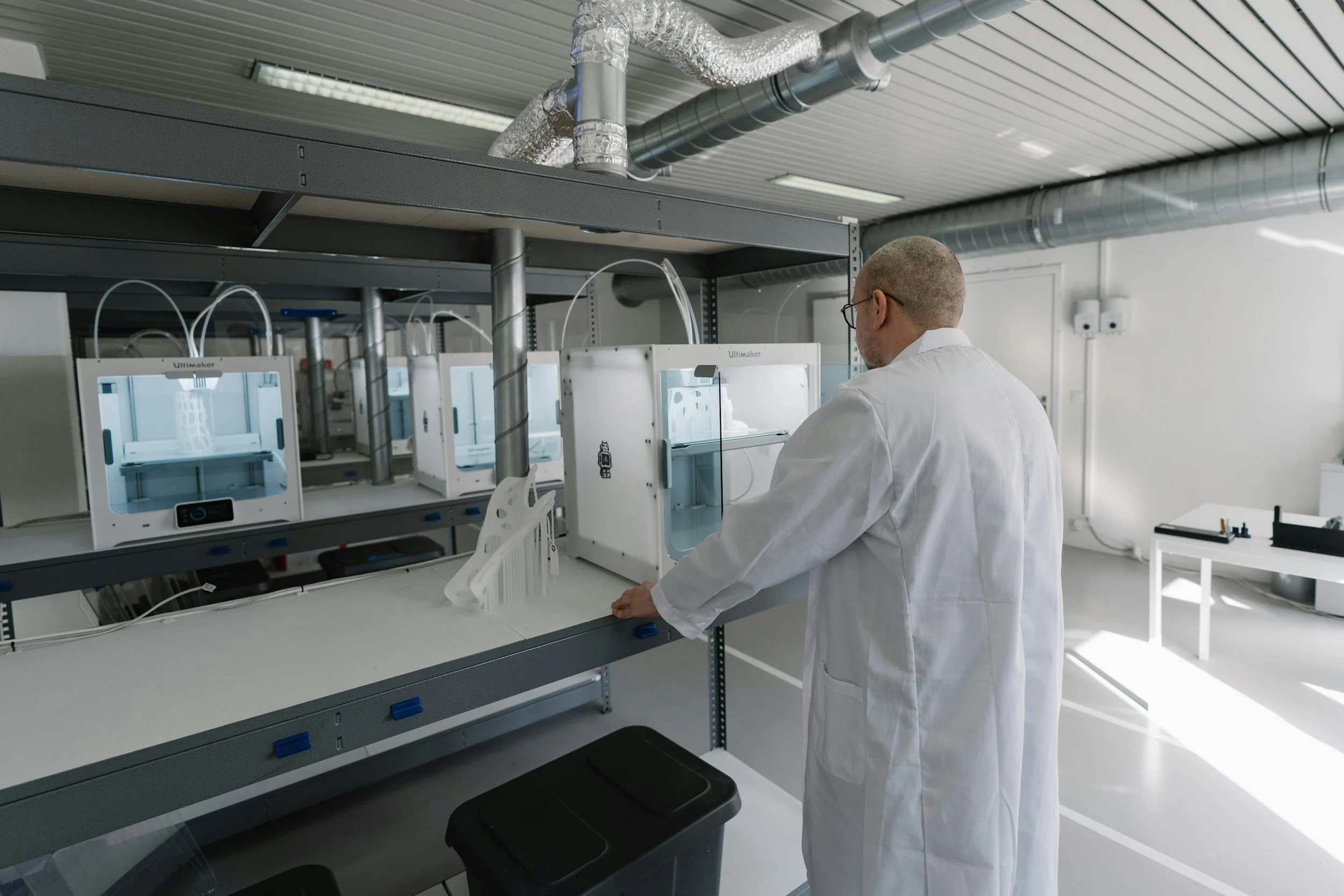 A scientist in a white lab coat working with 3D printers in a laboratory with ventilation ducts on the ceiling.