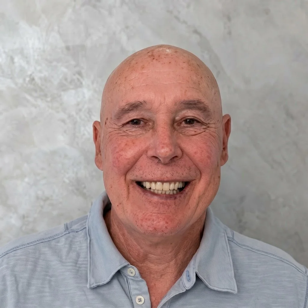 A smiling man with a bald head and freckles, wearing a light gray collared shirt, standing against a textured light gray background.