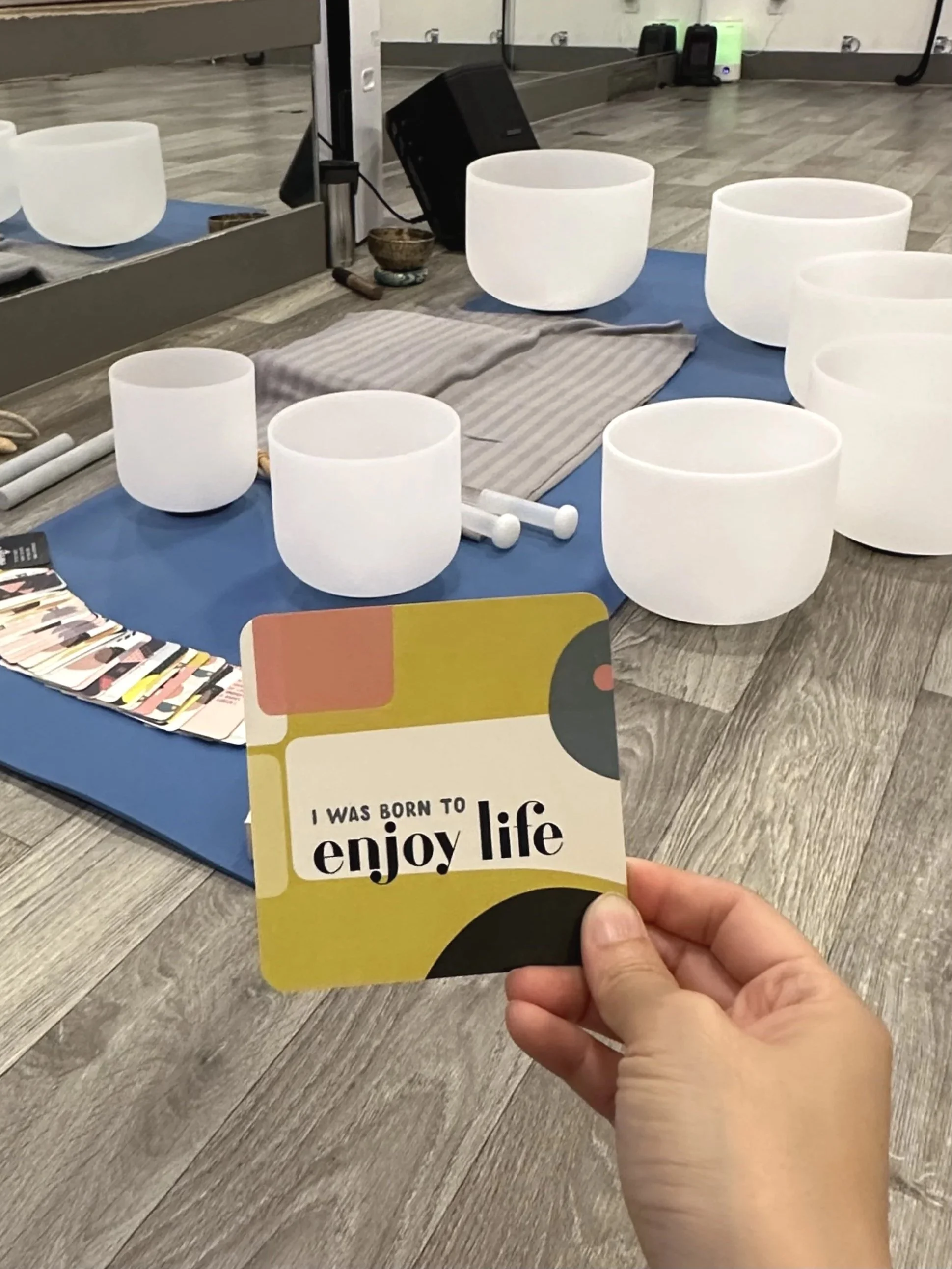 A hand holding a card that reads, 'I was born to enjoy life,' in front of a display with white crystal singing bowls and a mirror.