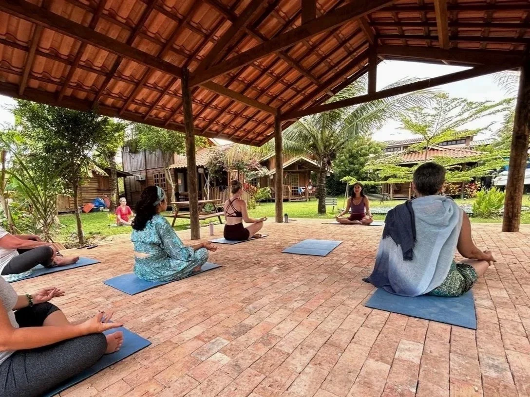 People practicing yoga outdoors under a wooden shelter, seated on yoga mats on a brick floor, with trees and traditional wooden buildings in the background.