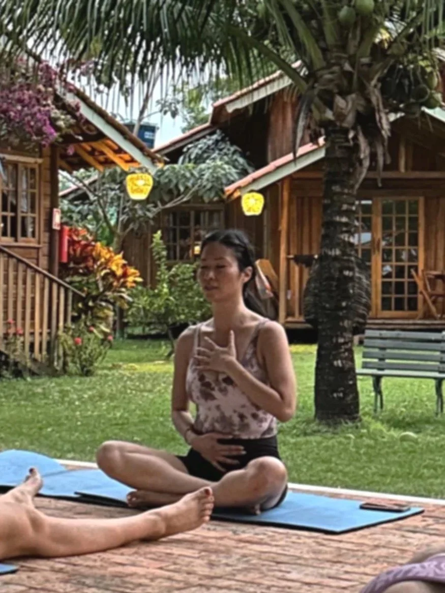 A woman practicing yoga outdoors in a tropical setting with wooden huts, palm trees, and flowering plants around her.